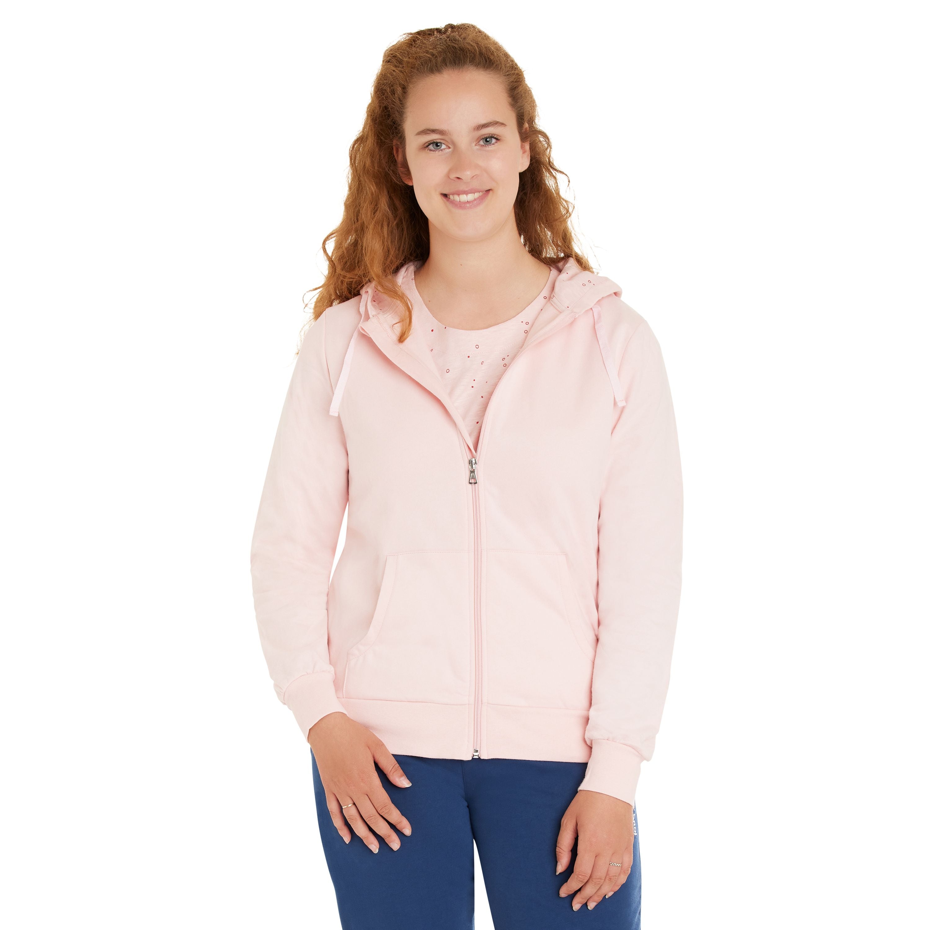 Eine lächelnde Frau mit langen lockigen Haaren, die einen hellrosa Kapuzenpulli mit Reißverschluss und eine blaue Hose trägt, steht mit dem Gesicht nach vorne vor einem schlichten weißen Hintergrund.