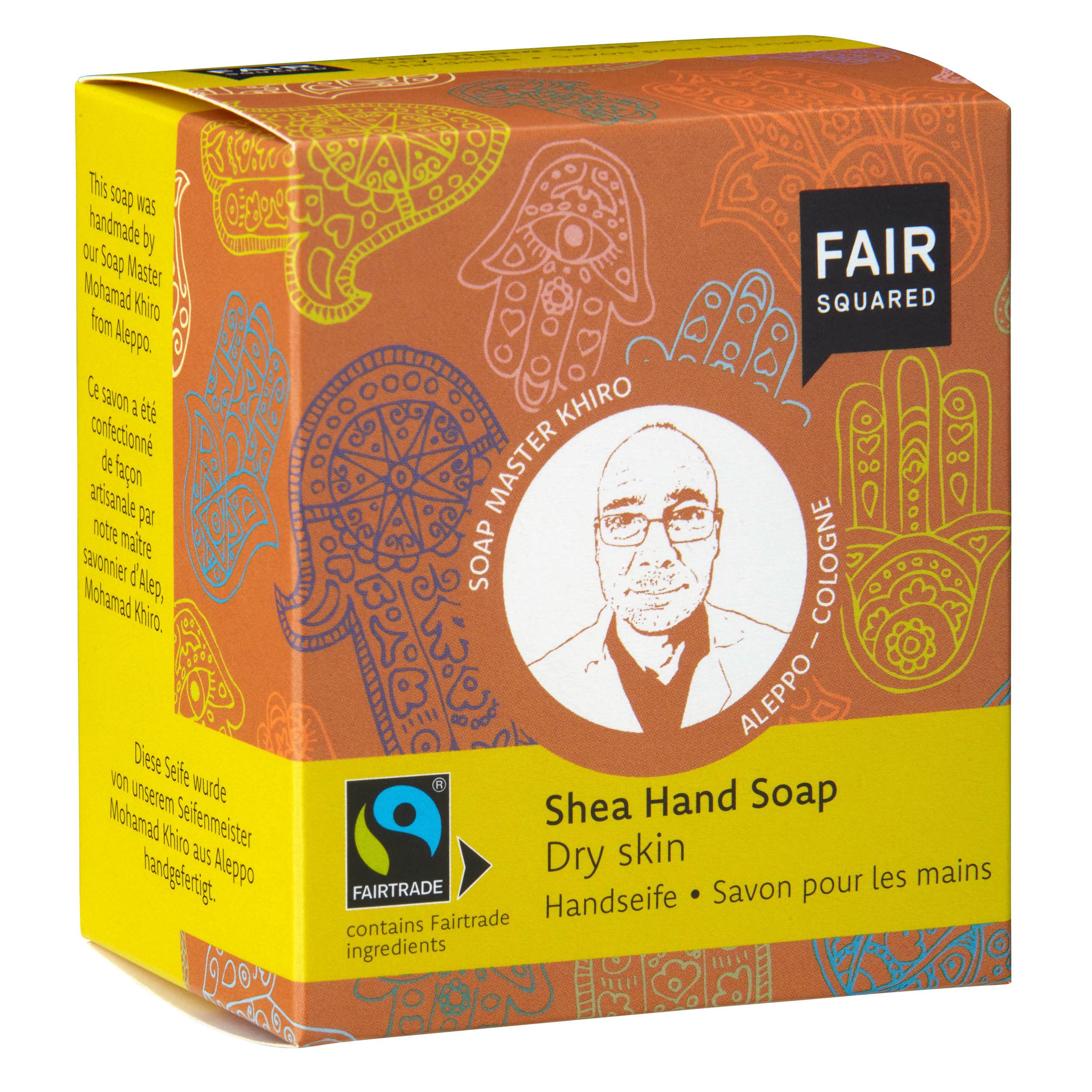 Eine Schachtel Fair Squared Shea-Handseife für trockene Haut mit einem illustrierten Porträt von Seifenmeister Khiro und bunten Hamsa-Motiven auf orangefarbenem Hintergrund. Auf der Schachtel stehen Fairtrade- und Produktinformationen in mehreren Sprachen.