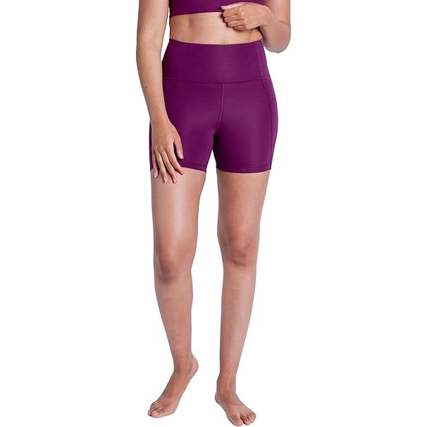 Eine Person, die einen lilafarbenen Sport-BH und hoch taillierte Sport-Shorts trägt und barfuß vor einem schlichten weißen Hintergrund steht. Ihre linke Hand ruht auf ihrer Taille.