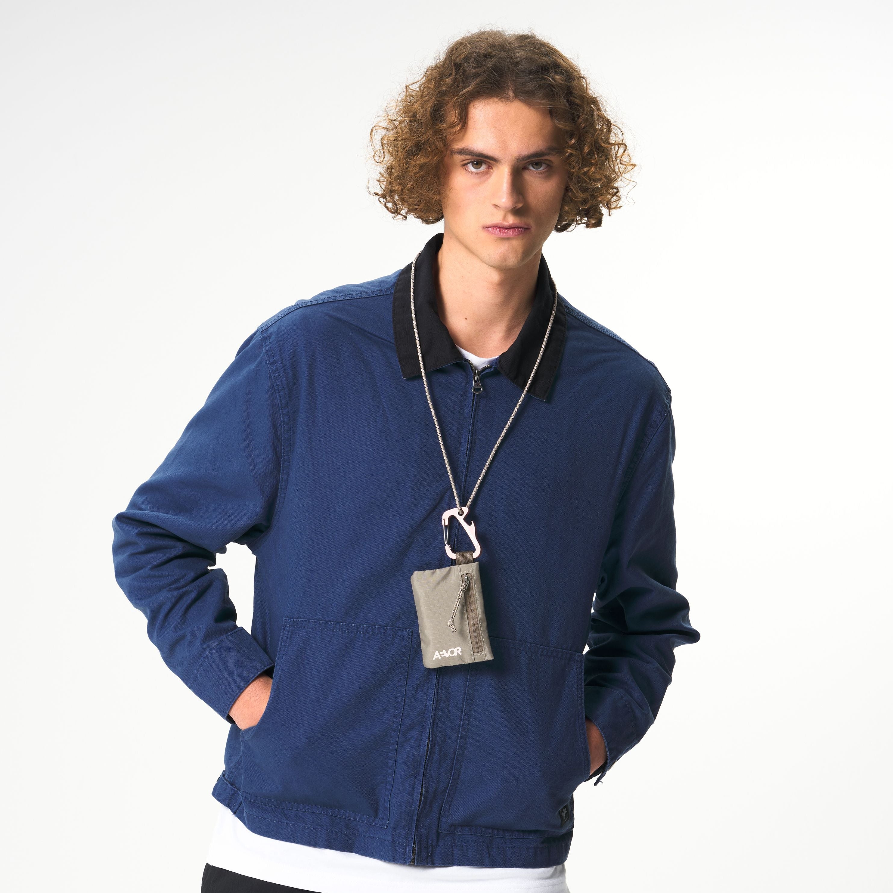 Eine junge Person mit lockigem Haar trägt eine blaue Jacke, ein weißes Hemd und eine kleine graue Tasche an einem Schlüsselband um den Hals. Sie steht vor einem schlichten, hellen Hintergrund und hat die Hände in den Taschen.