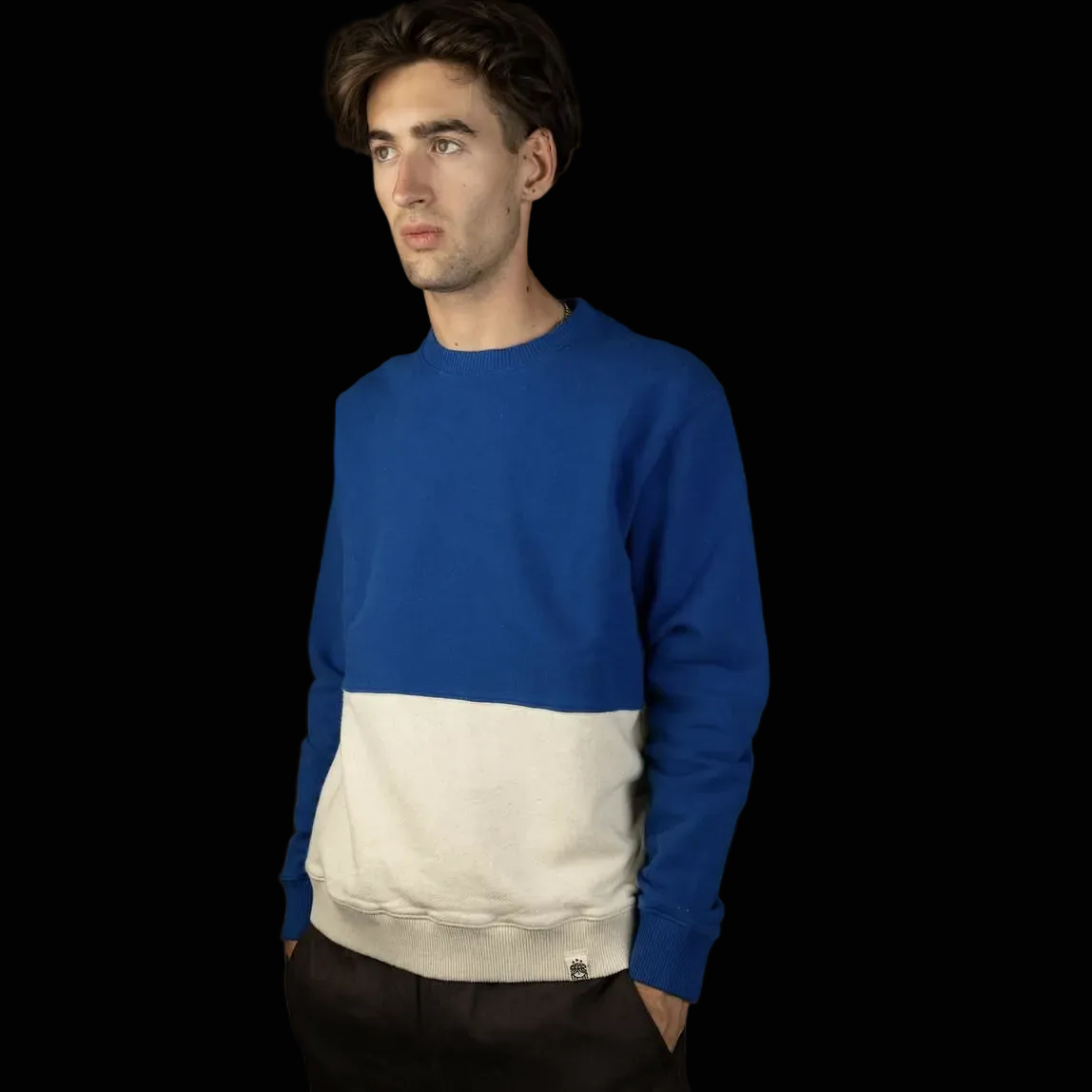 Ein Mann mit mittellangen braunen Haaren trägt ein blau-weißes Colorblock-Sweatshirt und eine dunkle Hose und steht vor einem schwarzen Hintergrund.