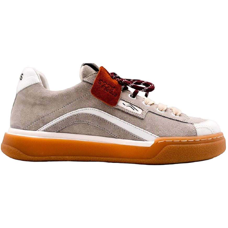 Ein beiger Wildleder-Sneaker mit weißen Akzenten, einer braunen Gummisohle, rot-schwarzen Schnürsenkeln und einem kleinen orangefarbenen Etikett in der Nähe der Schnürsenkel, dargestellt in einer Seitenansicht vor einem einfarbigen Hintergrund.