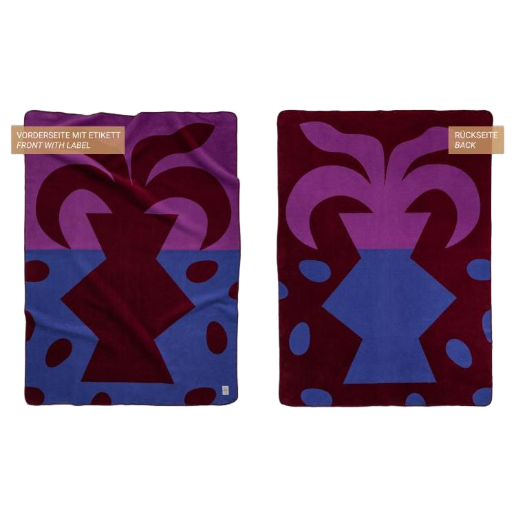 Zwei Ansichten einer rechteckigen Decke mit kühnen geometrischen Formen in Violett, Burgunderrot und Blau. Die linke Ansicht zeigt die Vorderseite mit einem kleinen Etikett, die rechte Ansicht zeigt die Rückseite ohne Etikett.