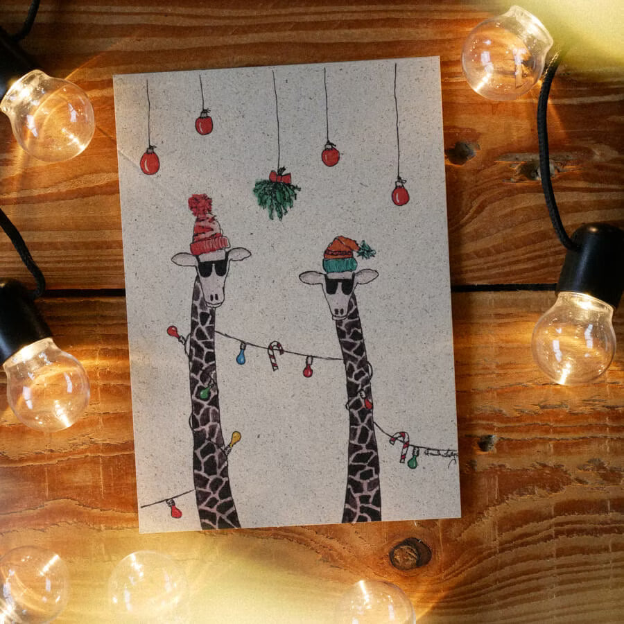 Weihnachtliche Karte mit zwei gezeichneten Giraffen in Mützen, umgeben von Lichterketten und weihnachtlicher Dekoration.