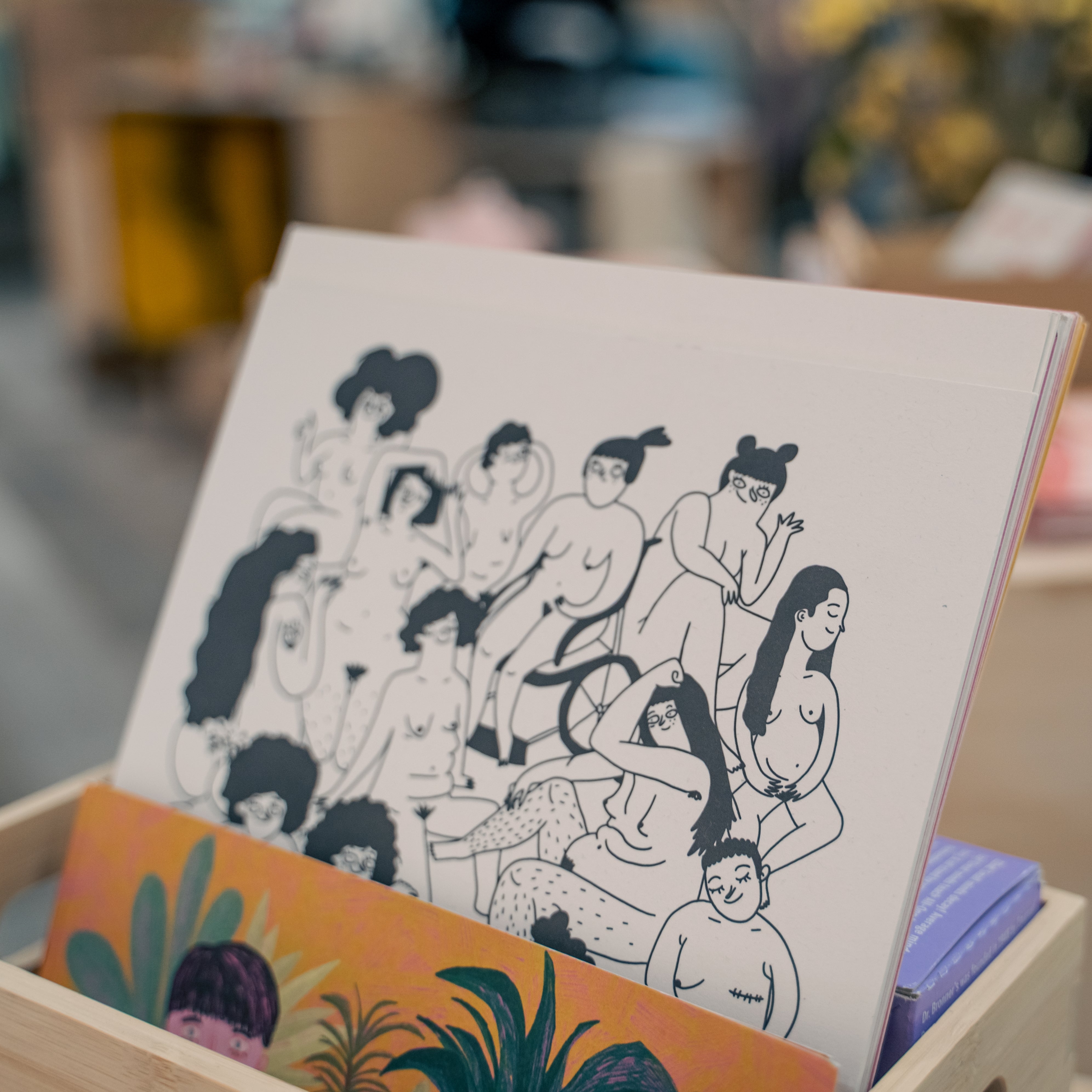 Ein Kunstbuch mit einer Zeichnung mehrerer nackter Menschen in einem Regal mit anderen bunten Büchern.