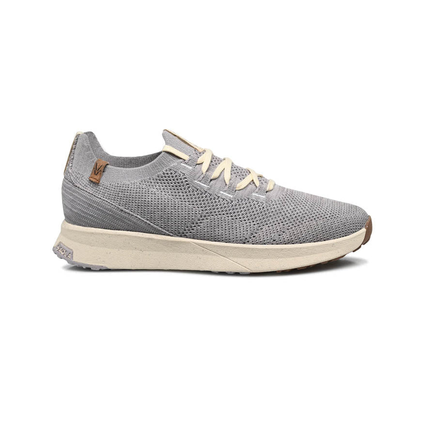 Ein grauer Strick-Sneaker mit beigen Schnürsenkeln und einer beigen Sohle, von der Seite vor weißem Hintergrund gezeigt. Der Schuh hat ein lässiges, leichtes Design und ein kleines braunes Logo-Tag in der Nähe der Ferse.