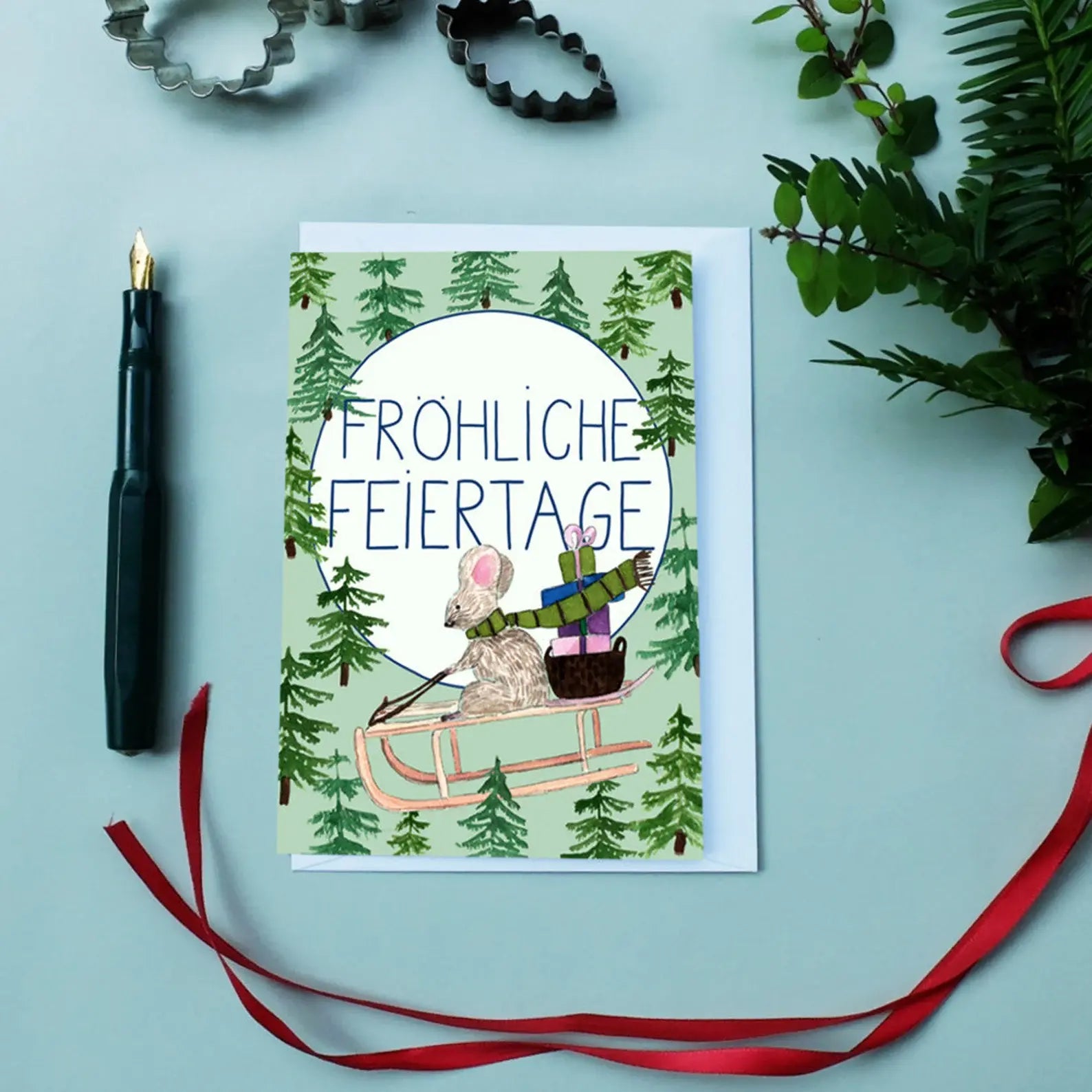 Weihnachtskarte mit Illustration einer Maus auf einem Schlitten, Text "Fröhliche Feiertage", daneben Stift und Dekoration.