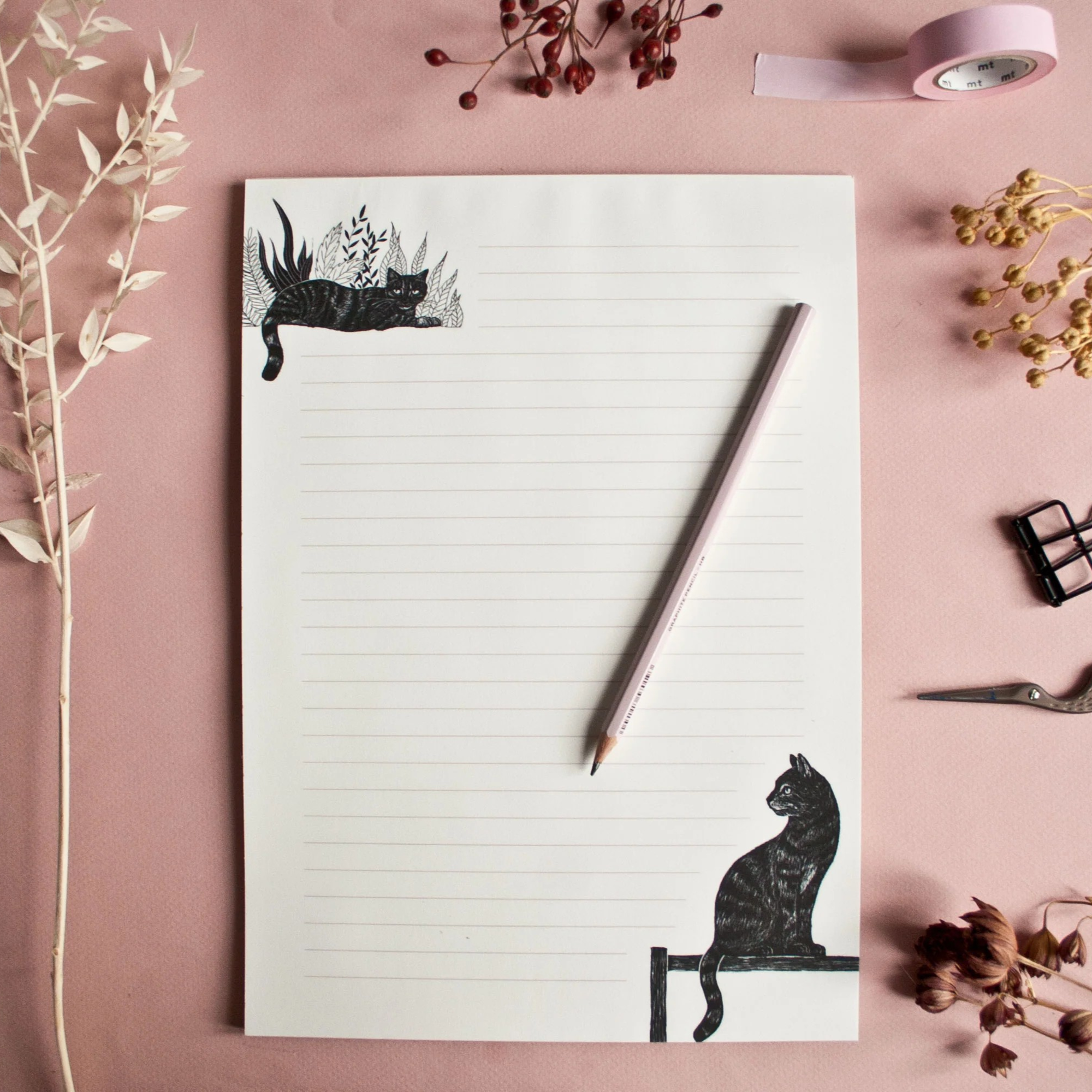 Schreibblock mit Katzenzeichnungen, einem Stift und dekorativen Pflanzen auf rosa Hintergrund.
