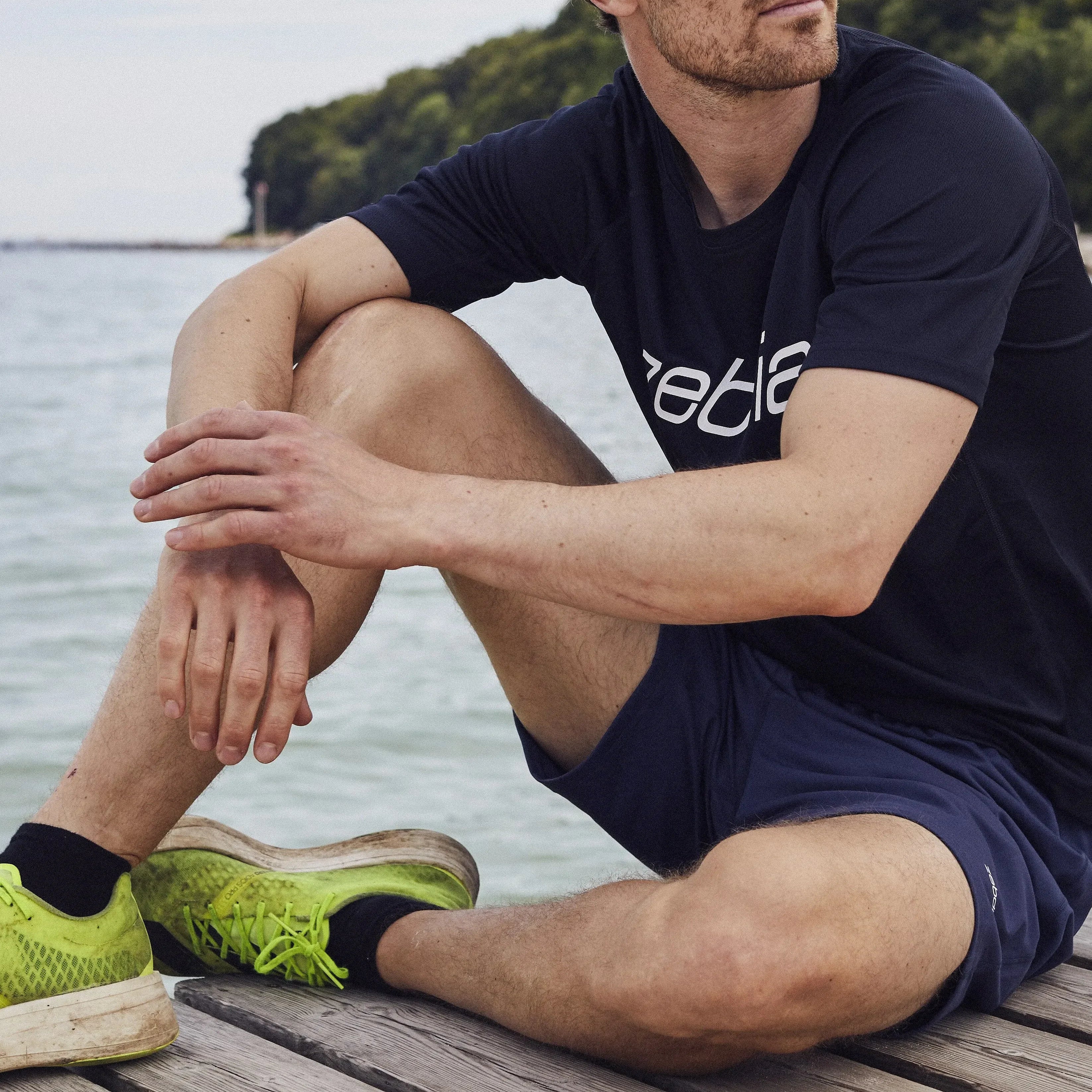 Ein Mann mit lockigem Haar und Bart sitzt auf einem Holzsteg am Wasser und trägt ein schwarzes T-Shirt, blaue Shorts und neongrüne Laufschuhe. Im Hintergrund sind Bäume und die Uferlinie zu sehen.