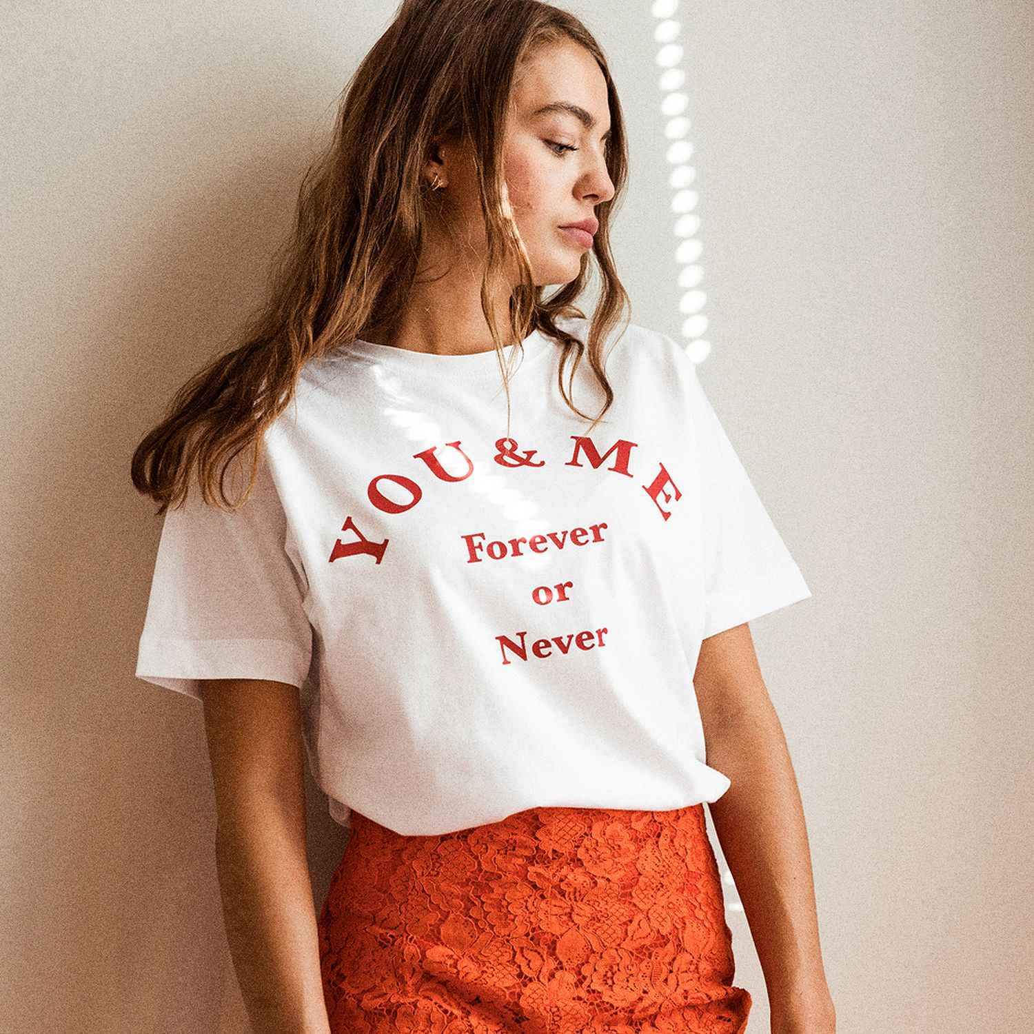 Frau in weißem T-Shirt mit rotem Text "YOU & ME Forever or Never" und einem roten Spitzenrock lehnt an einer Wand.