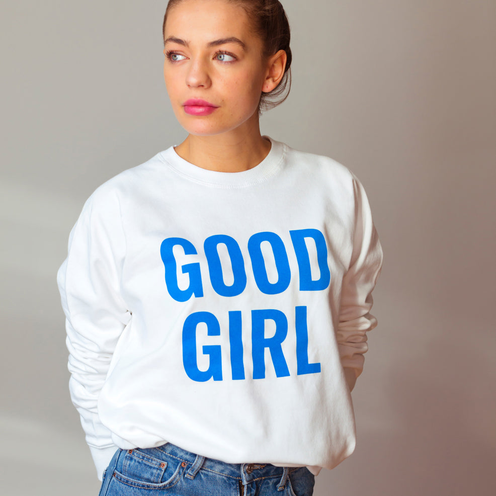 Frau in weißem Sweatshirt mit der Aufschrift "GOOD GIRL" in blauen Buchstaben, blickt zur Seite.