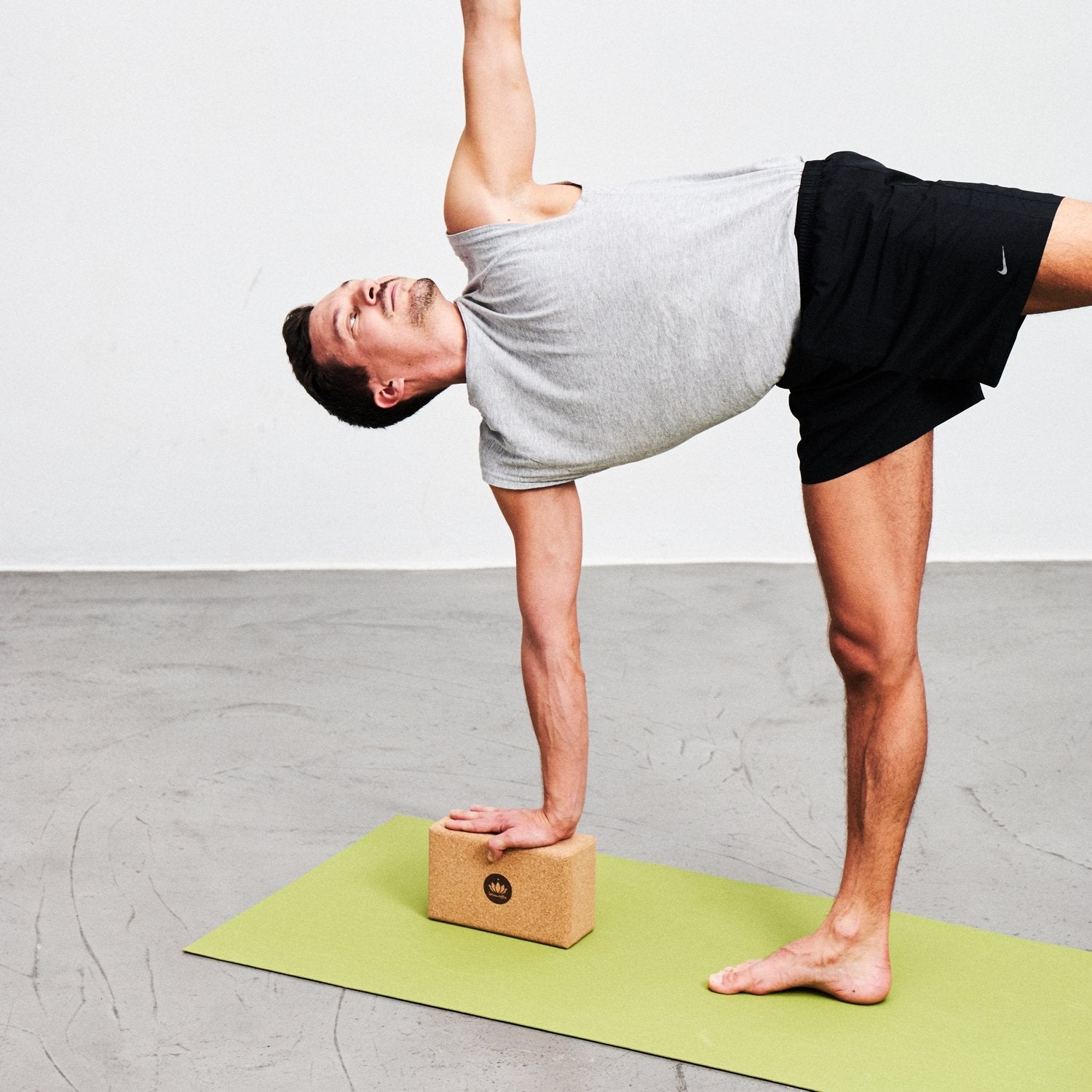 Mann macht Yoga auf einer grünen Matte, balanciert auf einem Fuß und stützt sich auf einem Holzblock ab.
