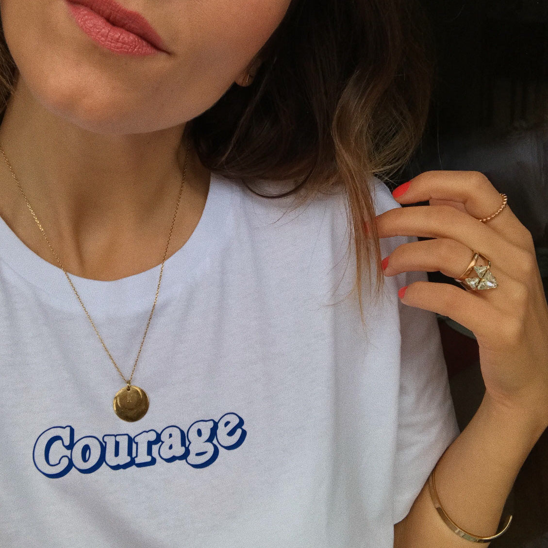 Frau trägt ein weißes T-Shirt mit der Aufschrift "Courage" und goldene Accessoires, einschließlich Halskette und Ringe.