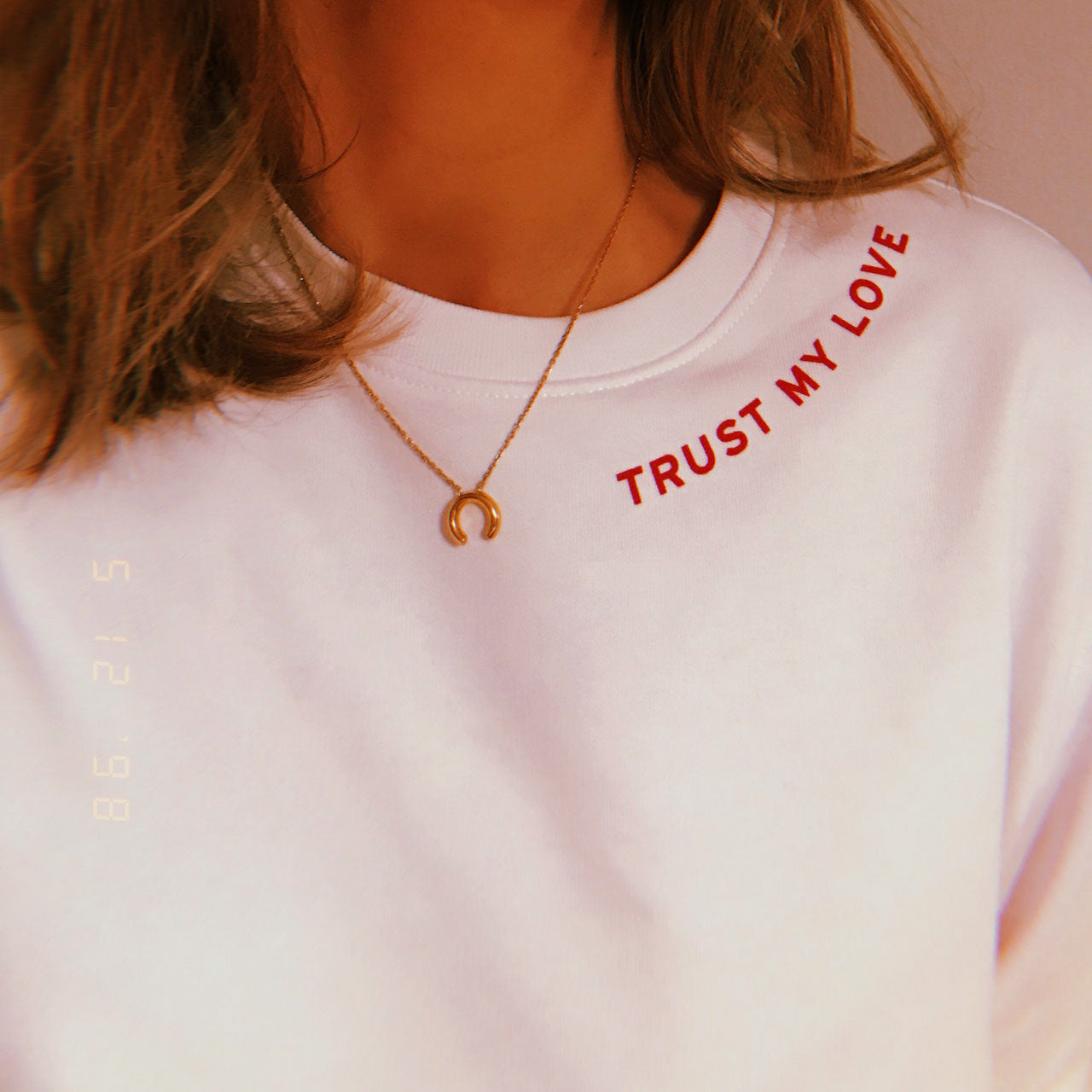 Frau trägt ein weißes Shirt mit rotem Schriftzug "TRUST MY LOVE" und einer goldenen Halskette mit Hufeisenanhänger.