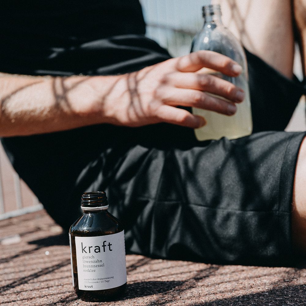 Person auf Boden sitzend, hält klare Flasche; daneben eine kleine braune Flasche mit Etikett "kraft".