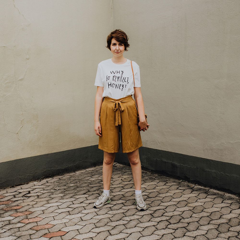 Eine Person steht in einem Innenhof und trägt ein weißes T-Shirt mit der Aufschrift „Why so perfect, honey?“