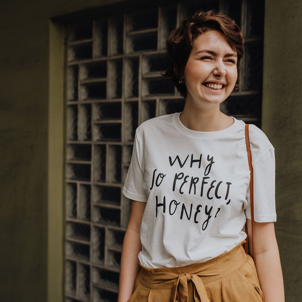 Lächelnde Person in weißem T-Shirt mit der Aufschrift "Why so perfect, honey?" vor einer grauen Wand.