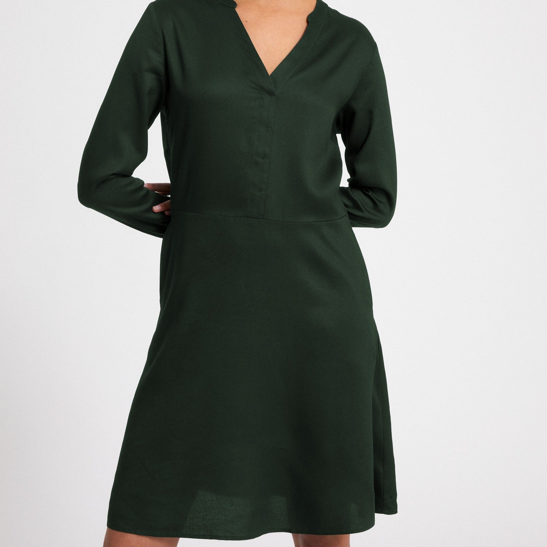 Person trägt ein dunkelgrünes langärmeliges Kleid mit V-Ausschnitt vor weißem Hintergrund.