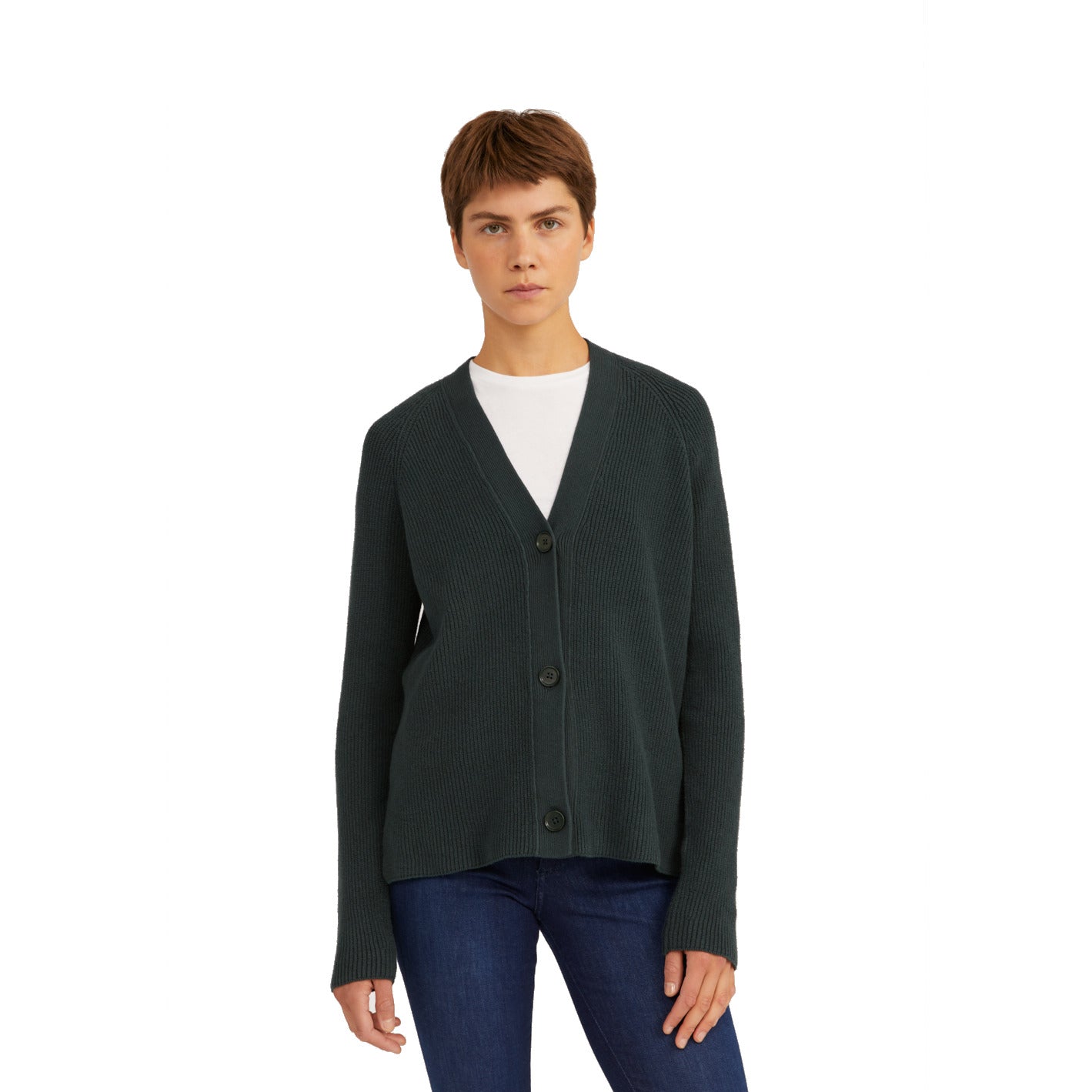 Person trägt eine dunkelgrüne Strickjacke über einem weißen T-Shirt und dunkelblauen Jeans, steht vor weißem Hintergrund.