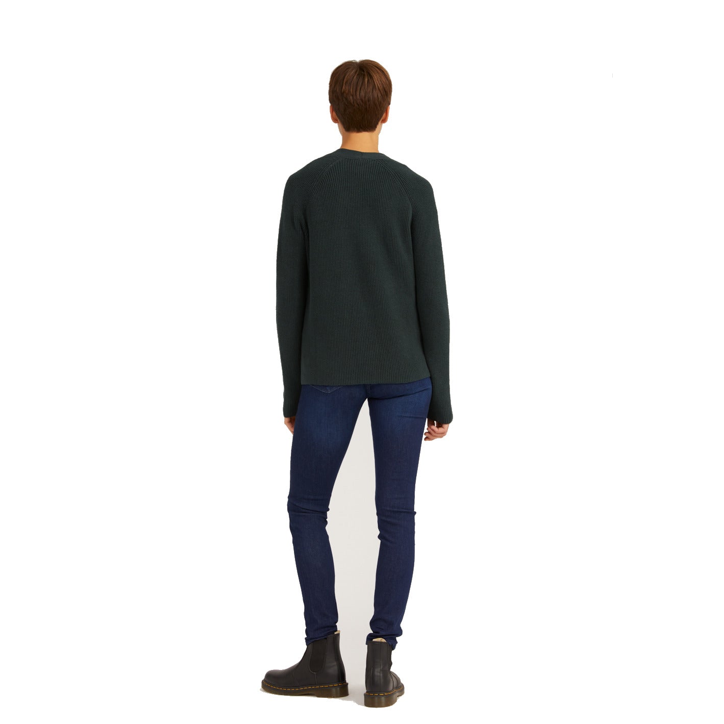 Person in dunkelgrünem Pullover und blauer Jeans, von hinten gesehen, trägt schwarze Stiefel, steht vor weißem Hintergrund.