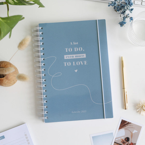 Blauer Kalender 2023 auf weißem Tisch, umgeben von einem goldenen Stift, Pflanzen und Fotos.