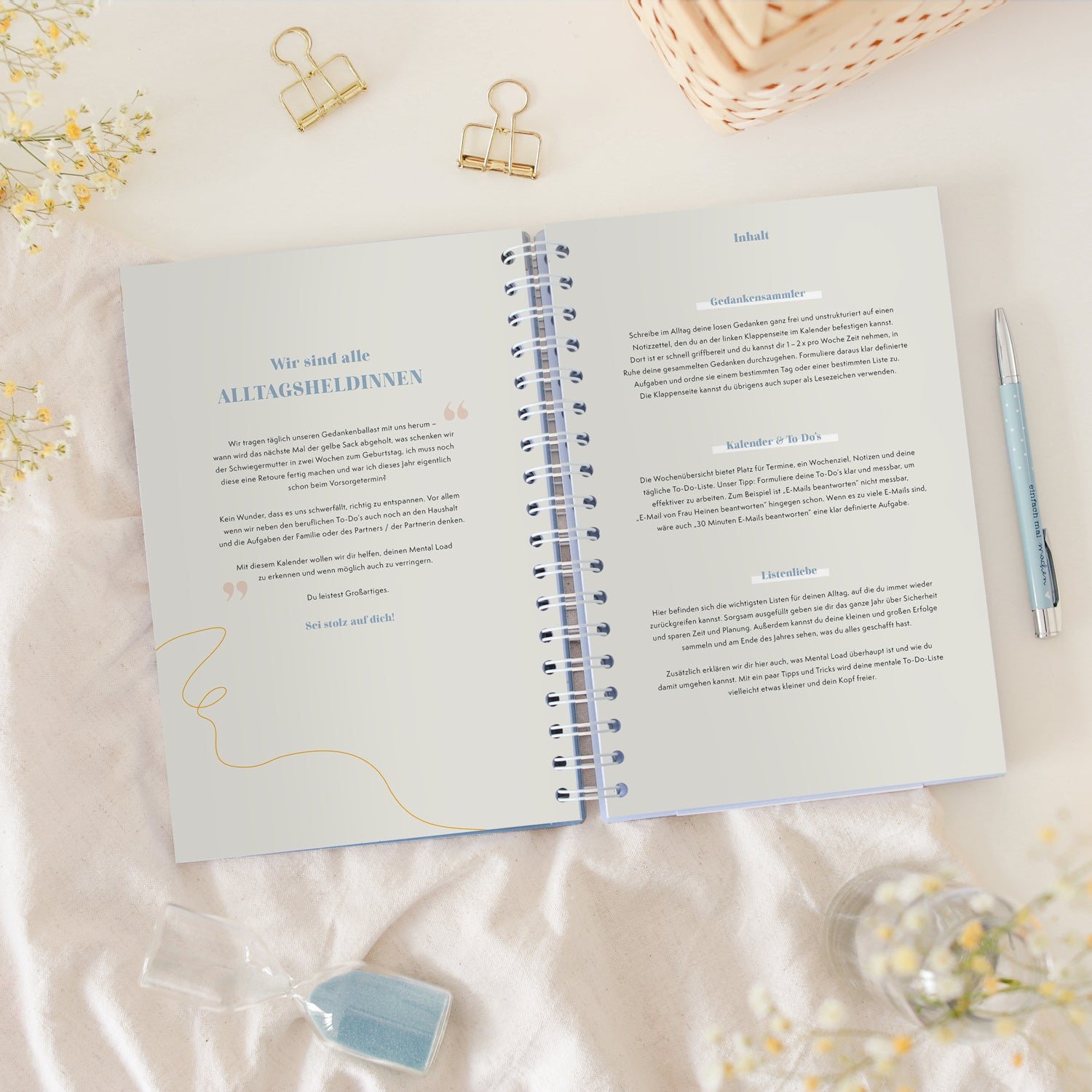 Aufgeschlagenes Notizbuch auf einem Tisch, umgeben von einer Sanduhr, einem Stift und Blumen, mit deutschen Texten.