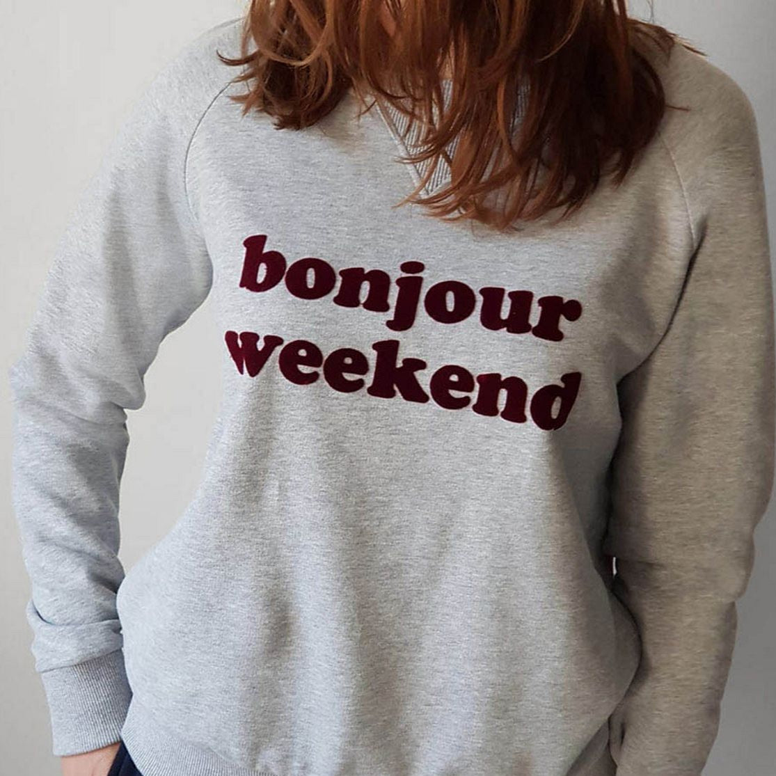 Person trägt einen grauen Pullover mit der Aufschrift "bonjour weekend" in dunkelroten Buchstaben.