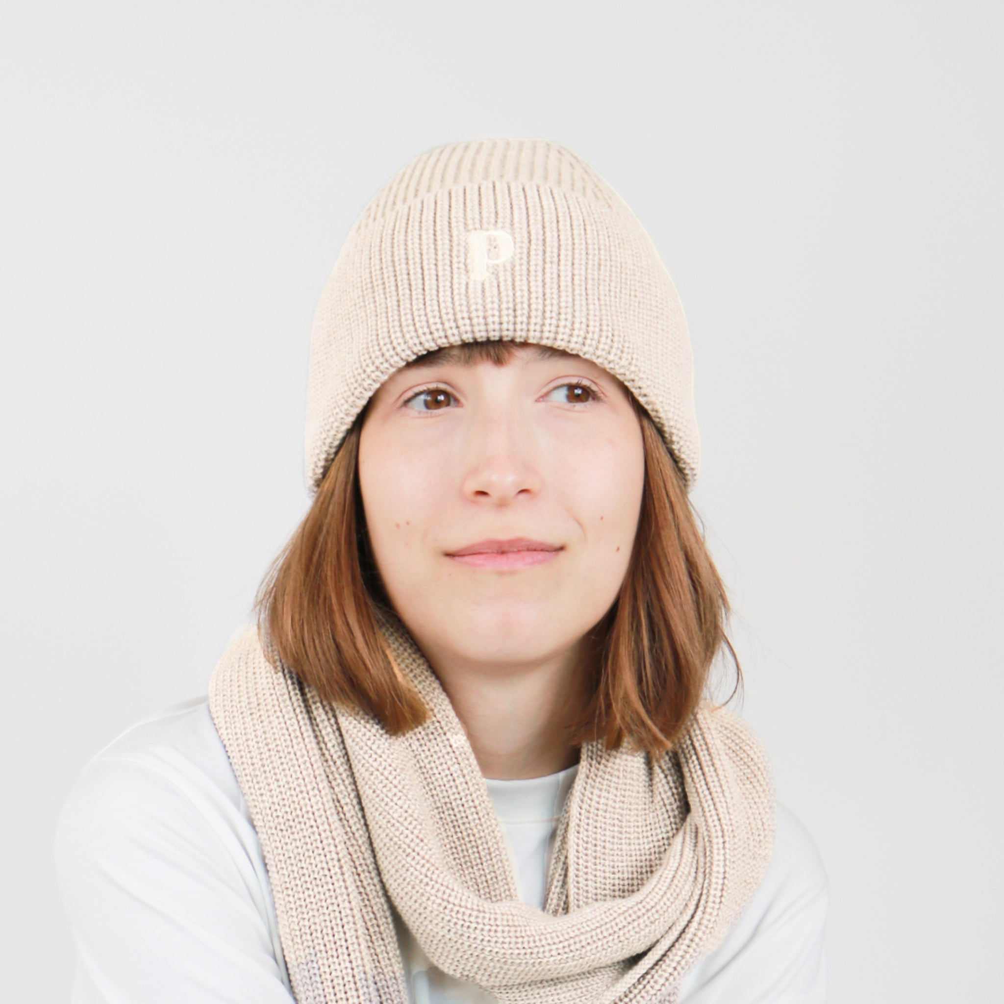 Frau mit beigefarbener Strickmütze und Schal schaut nach rechts vor hellem Hintergrund.