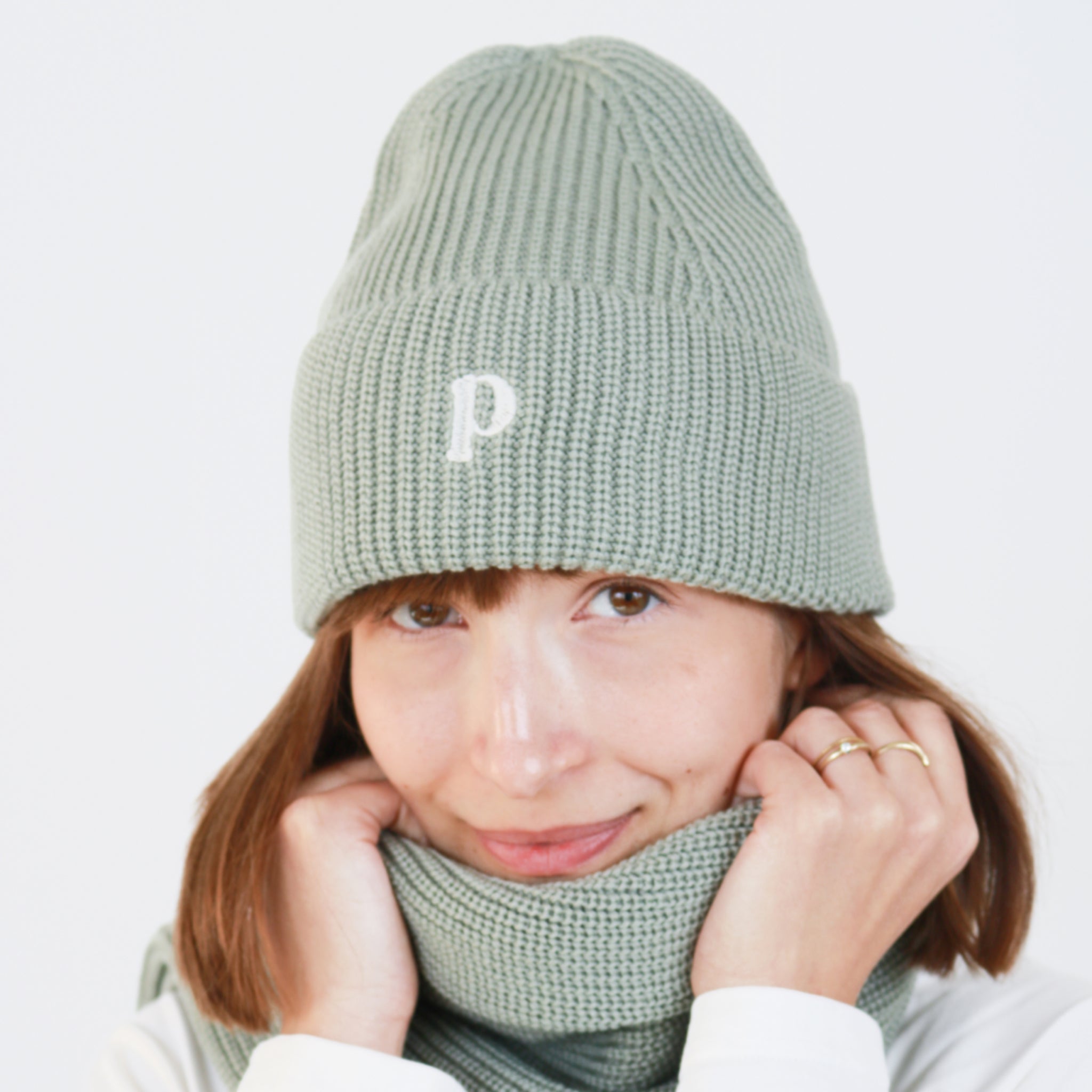 Person mit Strickmütze und Schal in hellem Grün, die Gesicht und Hände wärmt. Mütze mit eingesticktem Buchstaben "P".