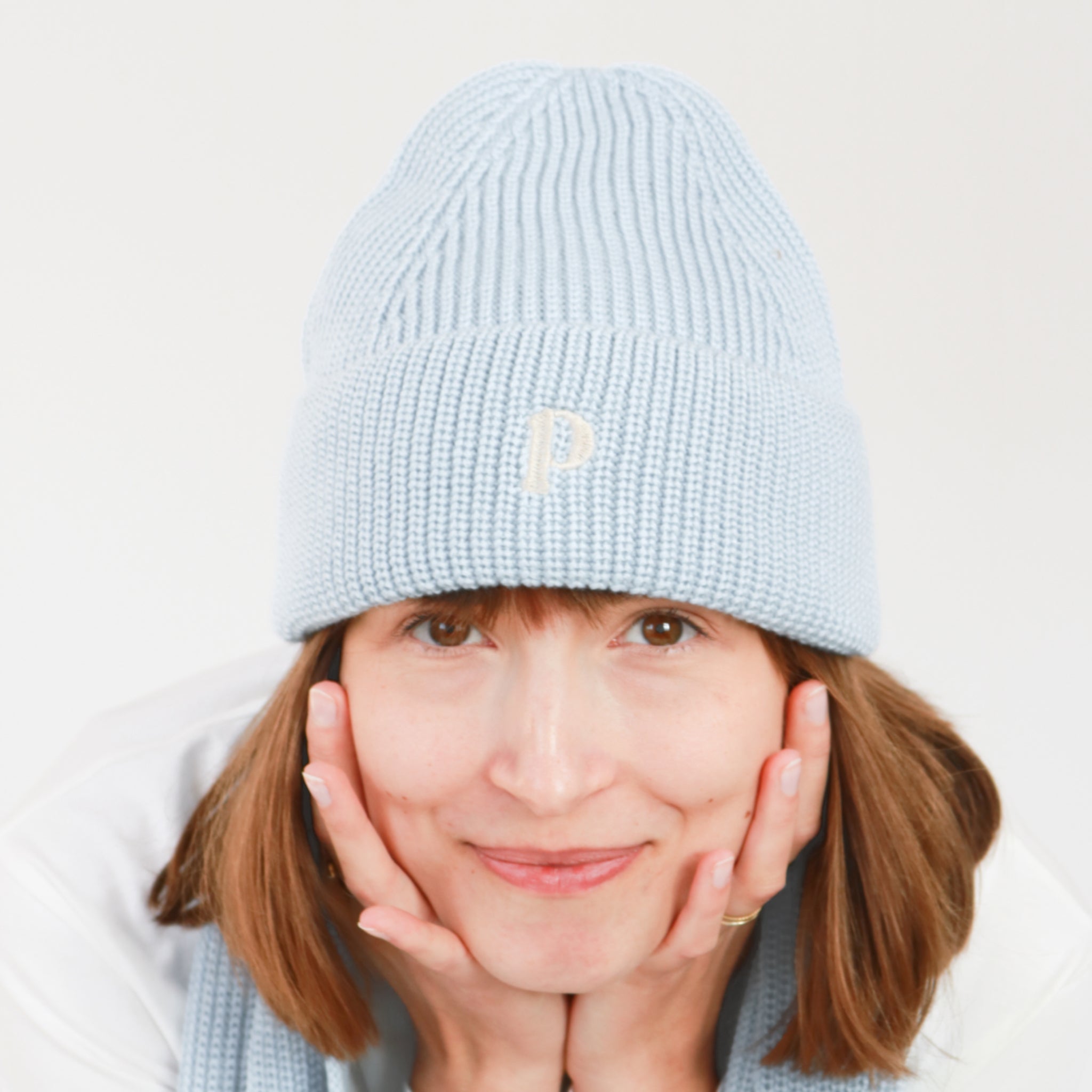 Frau mit blauem Strickmütze und Schal, stützt ihr Gesicht in die Hände und lächelt in die Kamera.