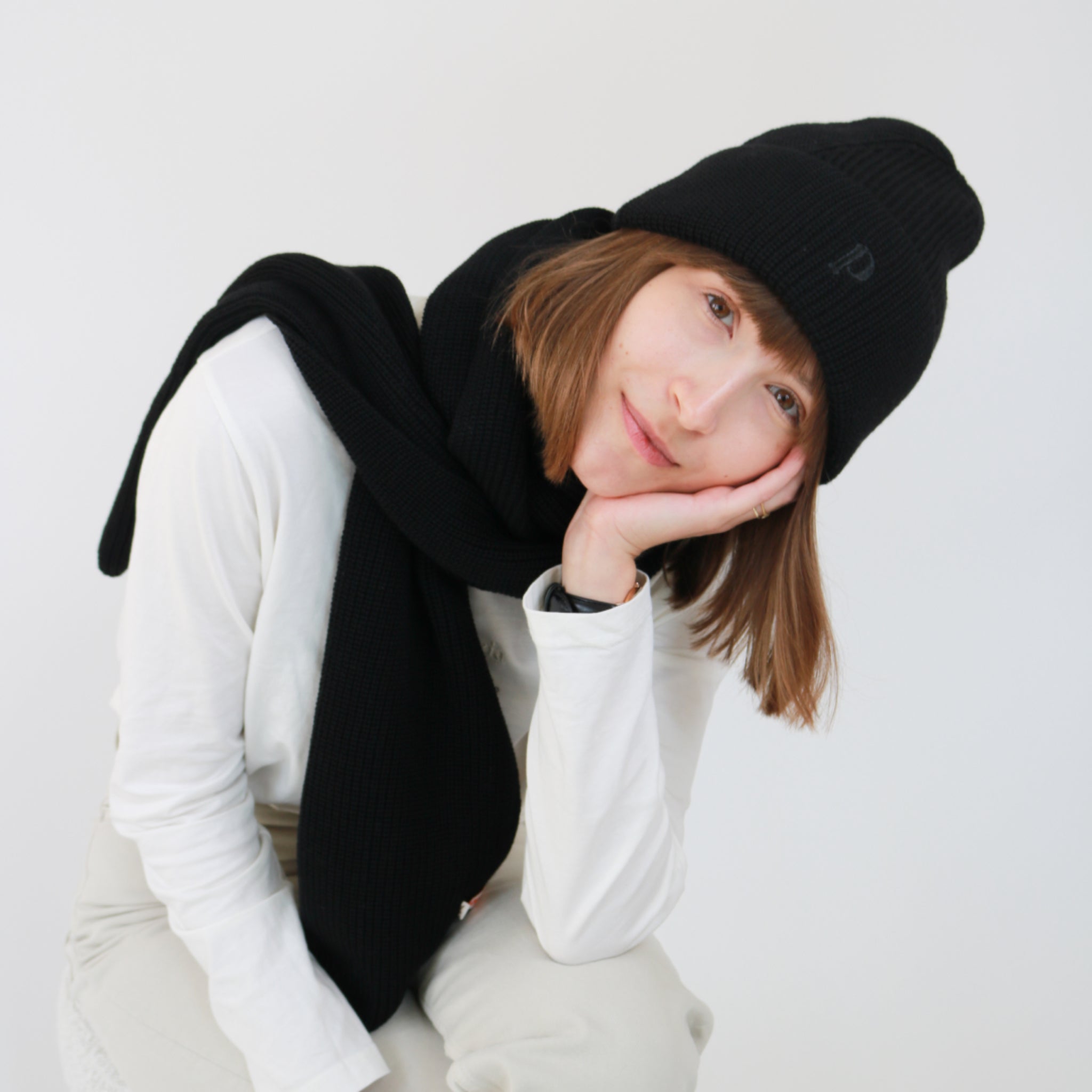 Person mit schwarzer Mütze und Schal in weißem Oberteil lächelt, während sie ihren Kopf auf die Hand stützt.