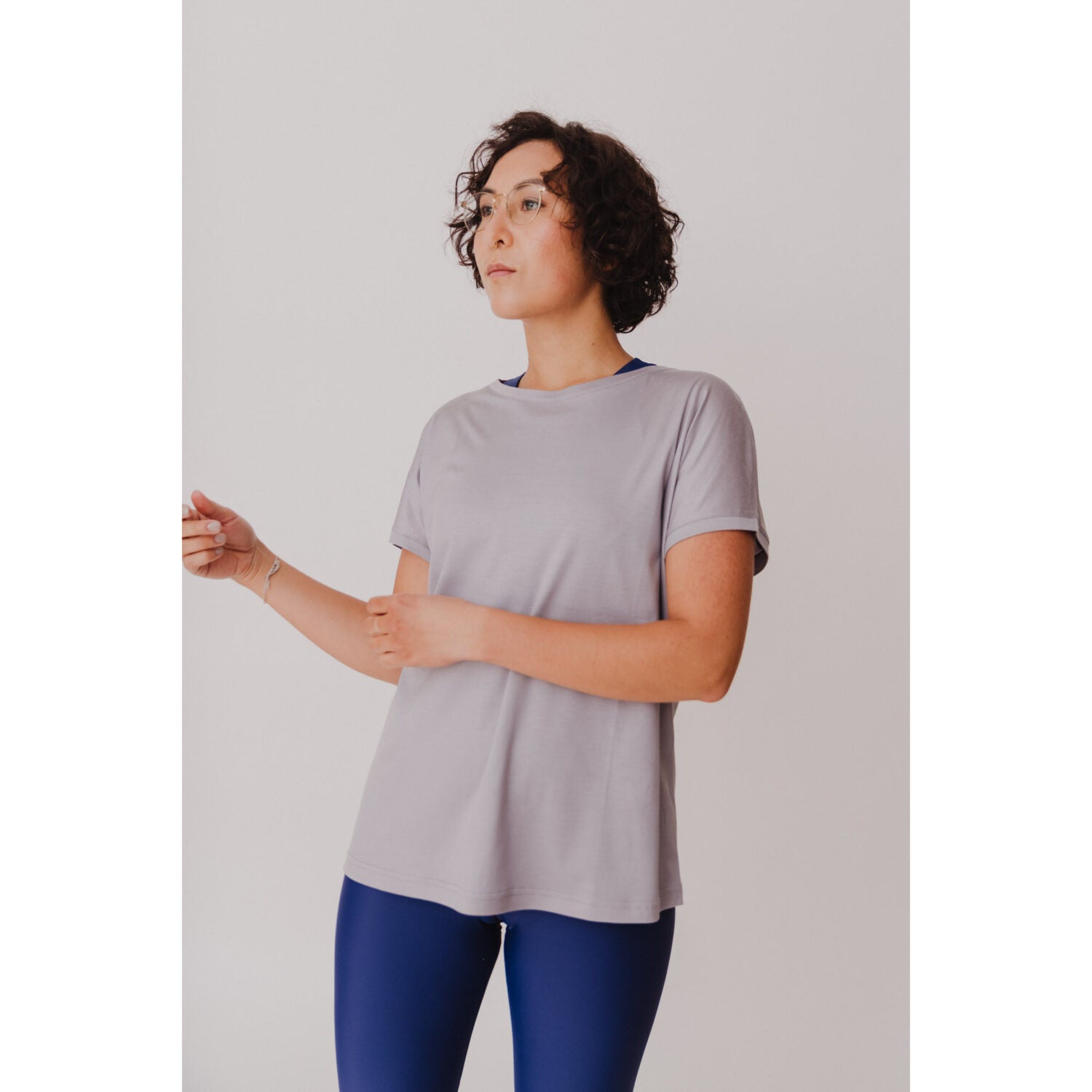 Person mit kurzen Locken trägt ein graues T-Shirt und blaue Leggings, stehend vor einem neutralen Hintergrund.