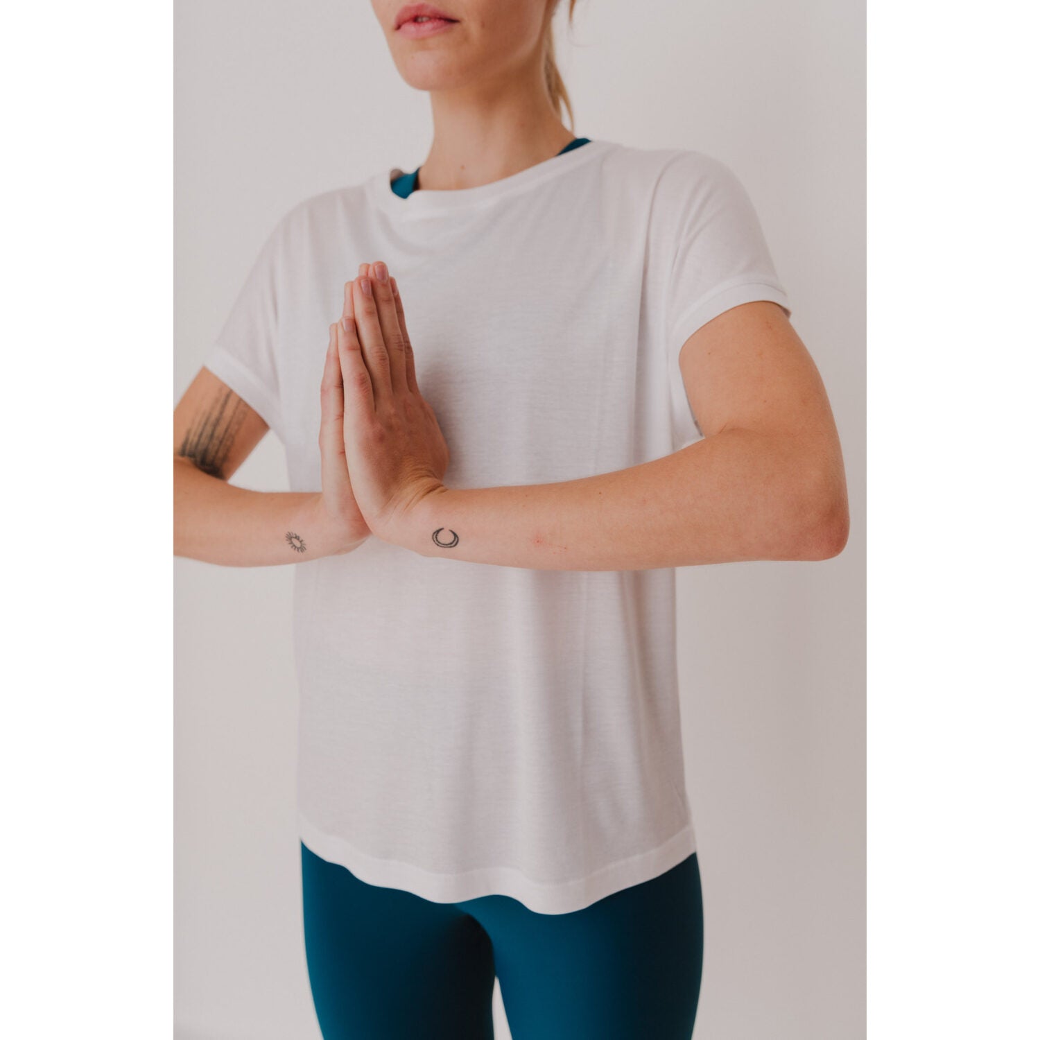 Person in weißem T-Shirt und blauer Leggings steht in Gebetshaltung, mit Handflächen zusammengeführt, in einem hellen Raum.