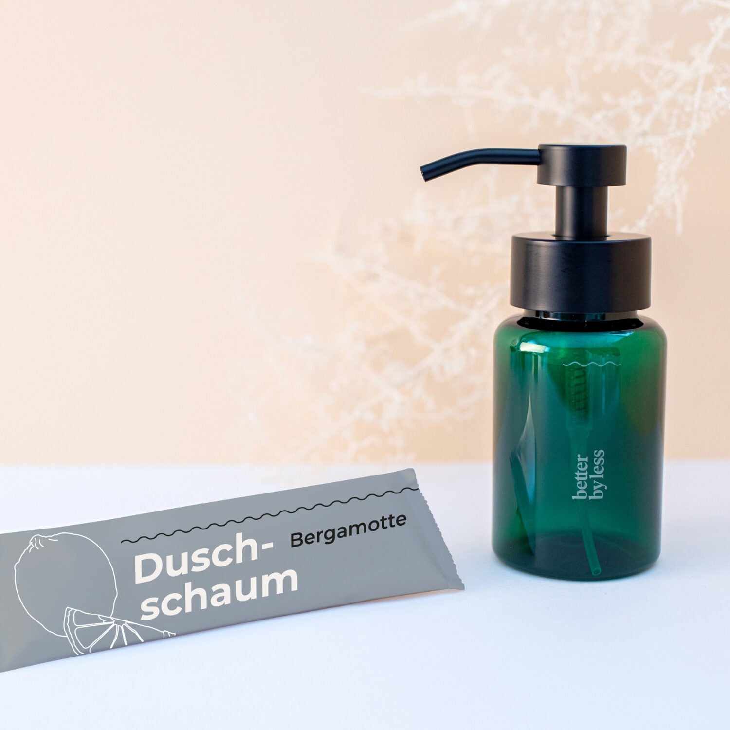 Grüne Flasche mit schwarzem Spender und graues Paket mit der Aufschrift "Duschschaum Bergamotte" auf hellem Hintergrund.