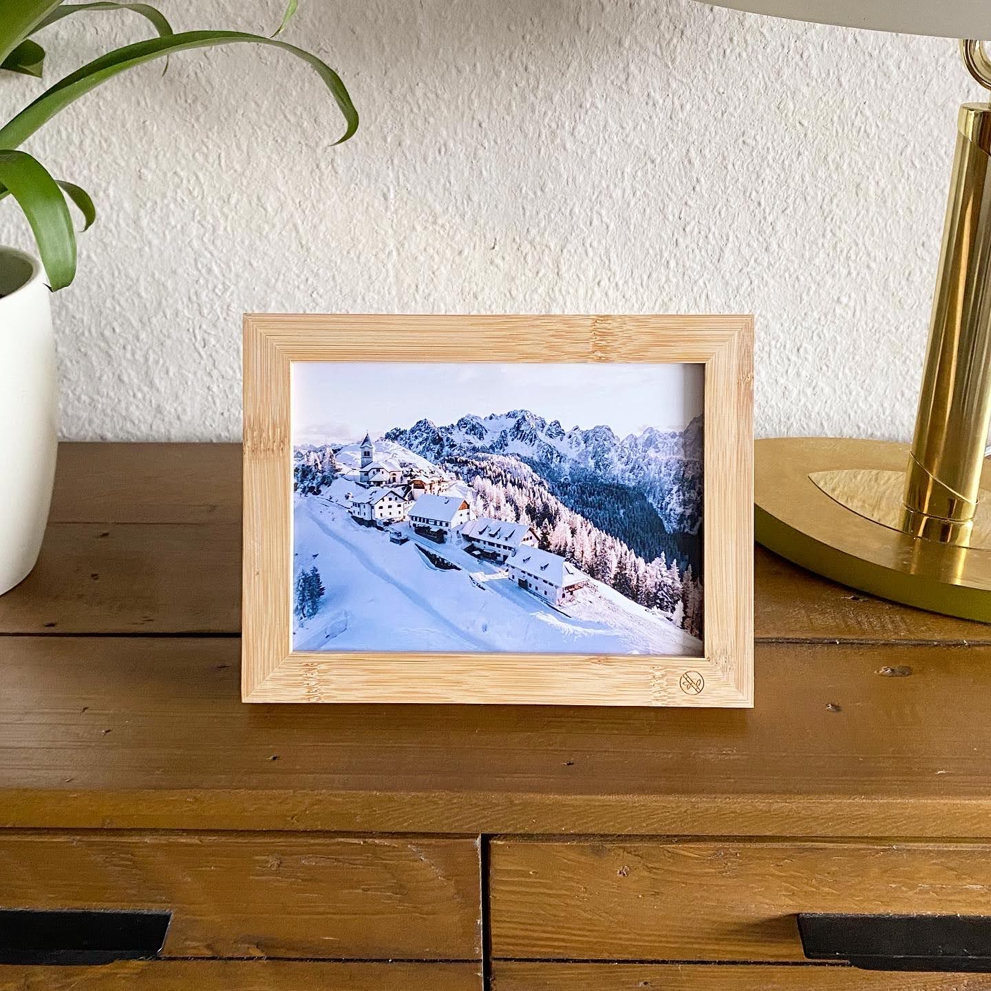 Gerahmtes Foto einer verschneiten Berglandschaft auf einem Holztisch neben einer Pflanze und einer goldenen Lampe.