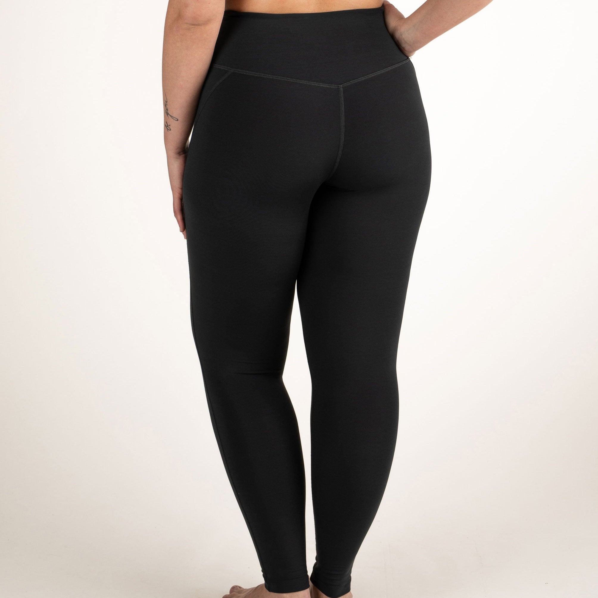 Frau in schwarzer Leggings von hinten, stehend vor neutralem Hintergrund.