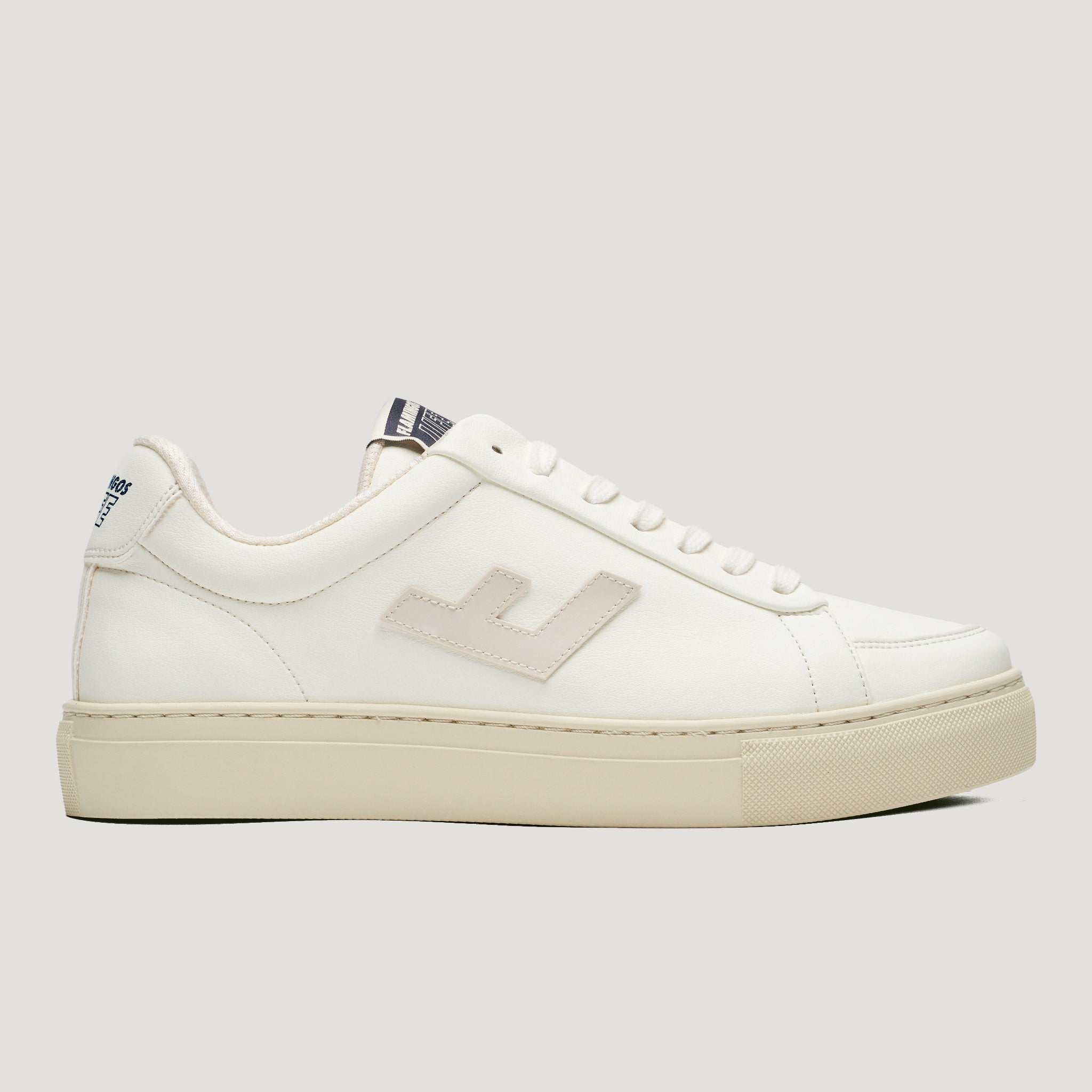 Weißer Sneaker mit beigem Emblem an der Seite, flachen Schnürsenkeln und einer cremefarbenen Sohle auf grauem Hintergrund.