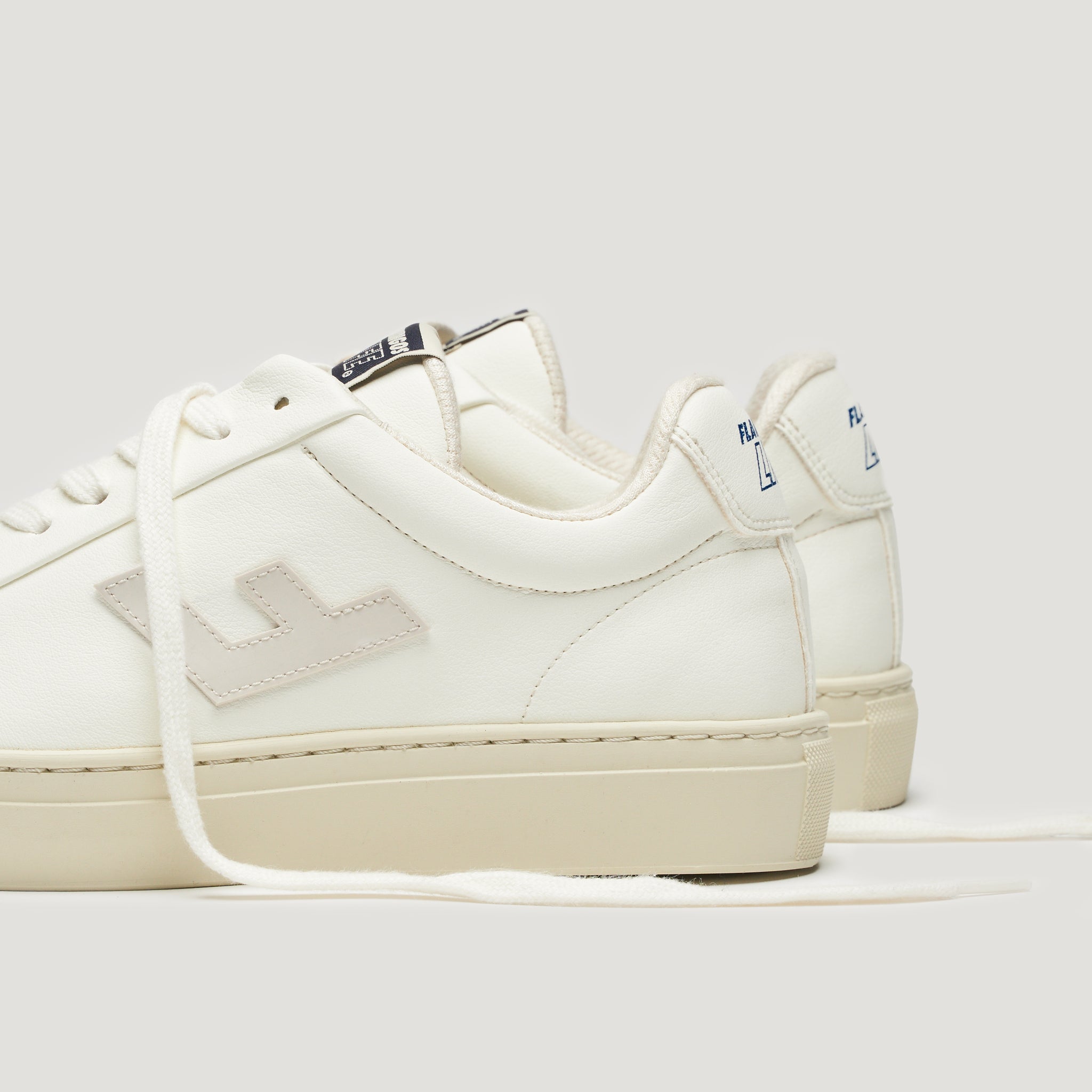 Weiße Sneaker mit beigem Logo und Sohlen auf grauem Hintergrund, einer davon mit sichtbarer Schnürung.