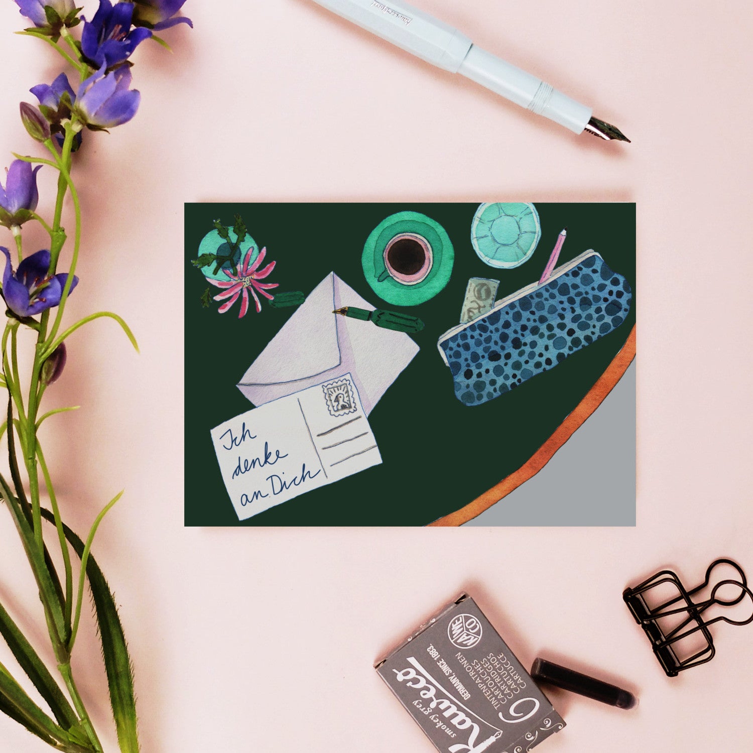 Postkarte mit der Aufschrift "Ich denke an Dich" neben einem Füller, Blumen, einer Maske und Büromaterial.