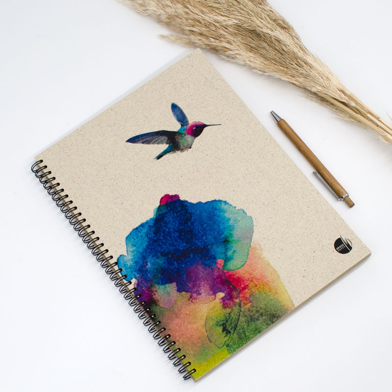 Notizbuch mit Aquarellkolibri und bunten Farbflecken, daneben ein Stift und trockene Pflanzen.