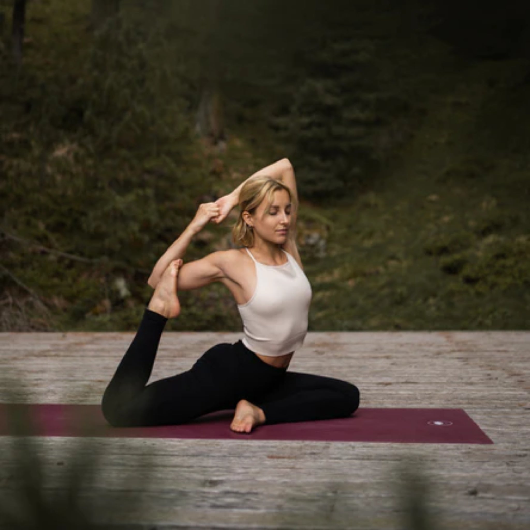 Frau in Yogapose auf lila Matte in einer waldigen Umgebung.