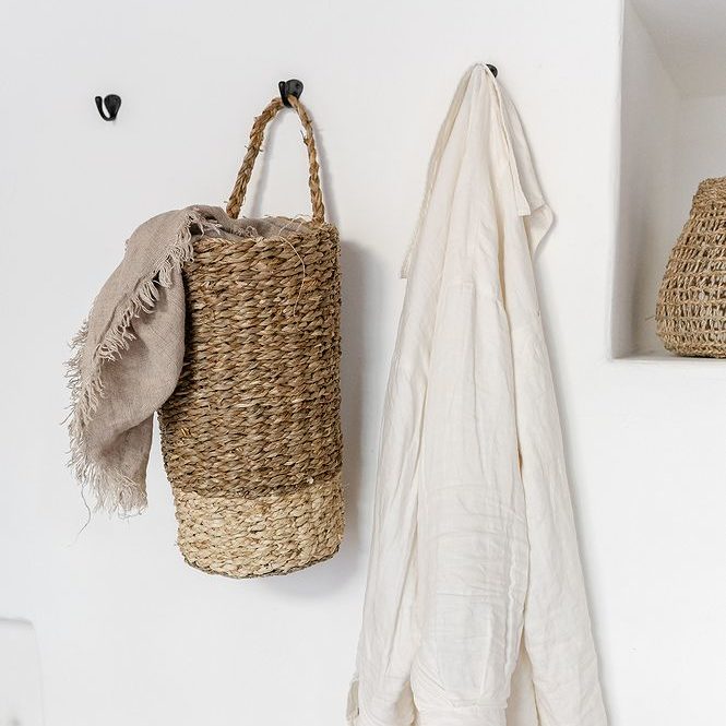 Geflochtener Korb mit beiger Decke und weißes Tuch an Wandhaken, vor weißem Hintergrund.