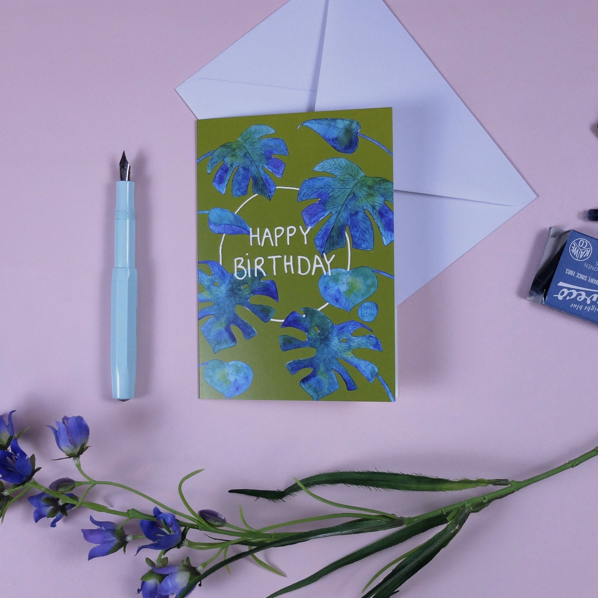 Geburtstagskarte mit blauen Blättern, daneben ein Füllfederhalter, ein Umschlag und violette Blumen auf rosa Hintergrund.