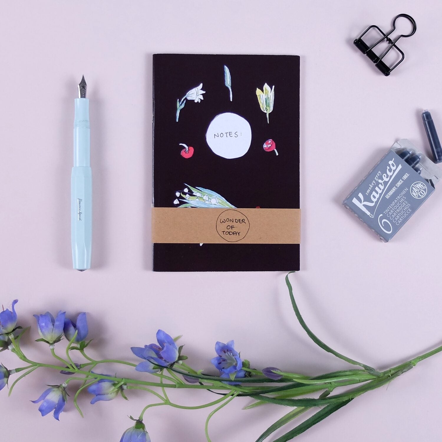 Notizbuch mit Blumen und Beeren, hellblauer Füller, Tintenpatronen und blauen Blumen auf rosa Hintergrund.