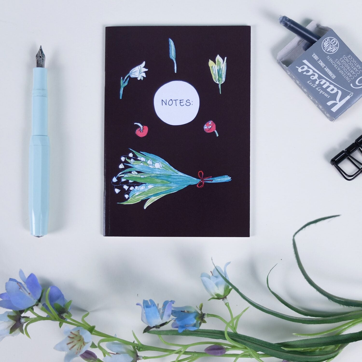 Notizbuch mit Blumenmotiven, einem blauen Füllfederhalter, Tinte und blauen künstlichen Blumen auf einem Tisch arrangiert.