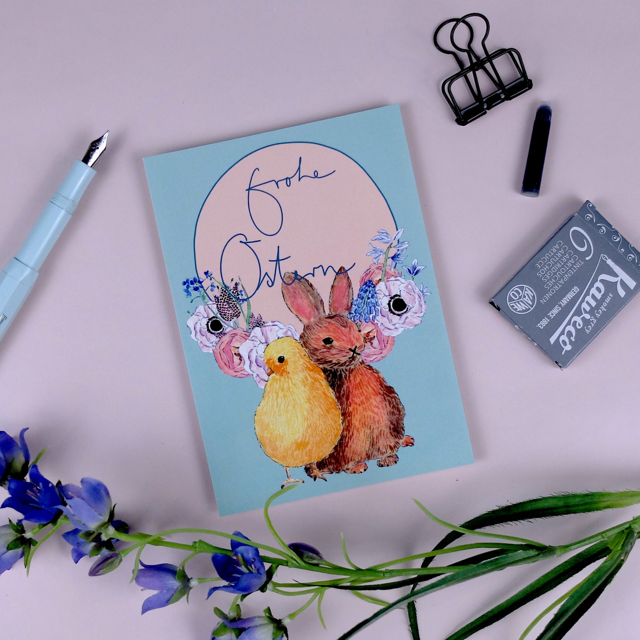Osterkarte mit Hase und Küken auf rosa Hintergrund, umgeben von einem Füller, einer Klammer und lila Blumen.