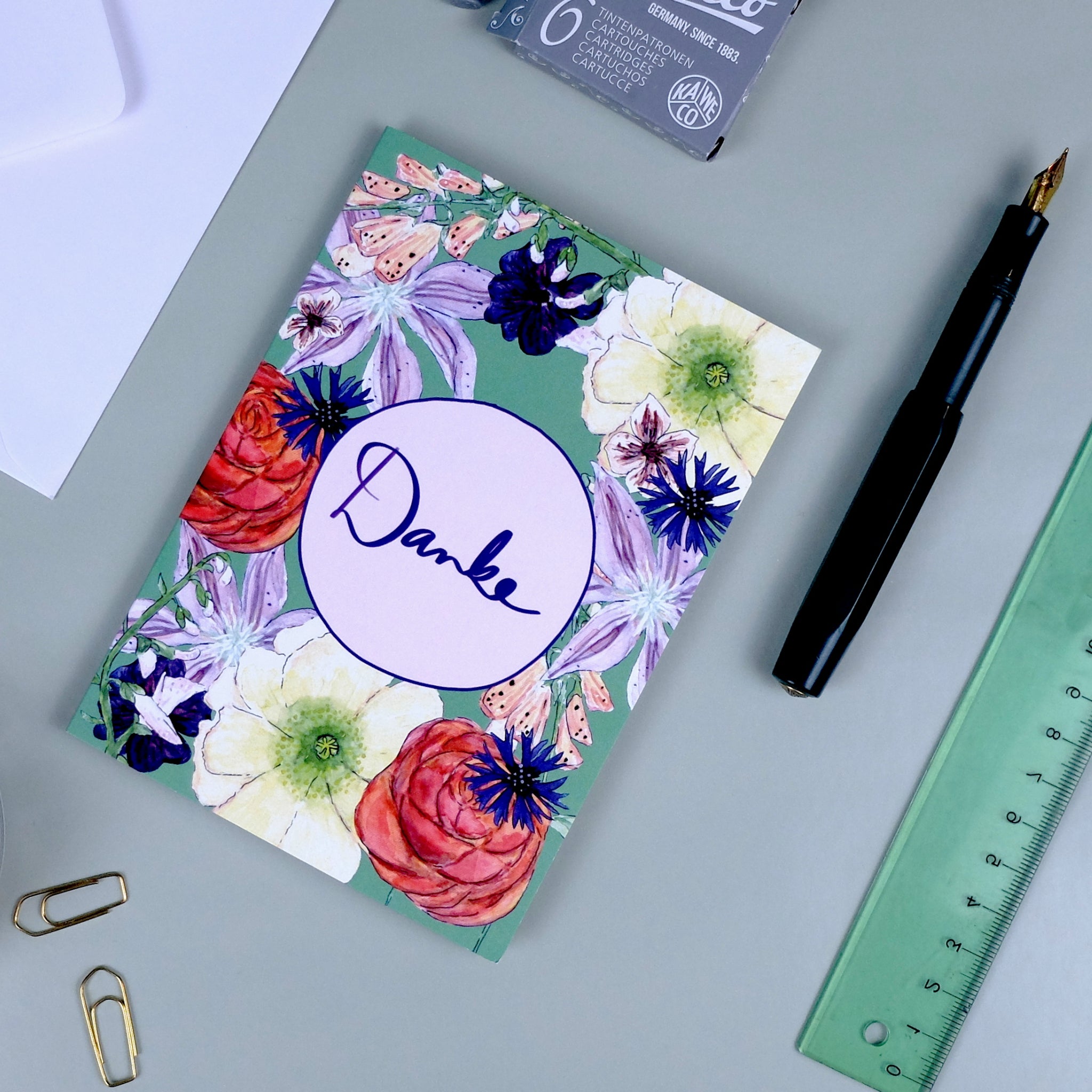 Grußkarte mit "Danke" und Blumendesign liegt auf Schreibtisch neben Füller, Lineal und Büroklammern.