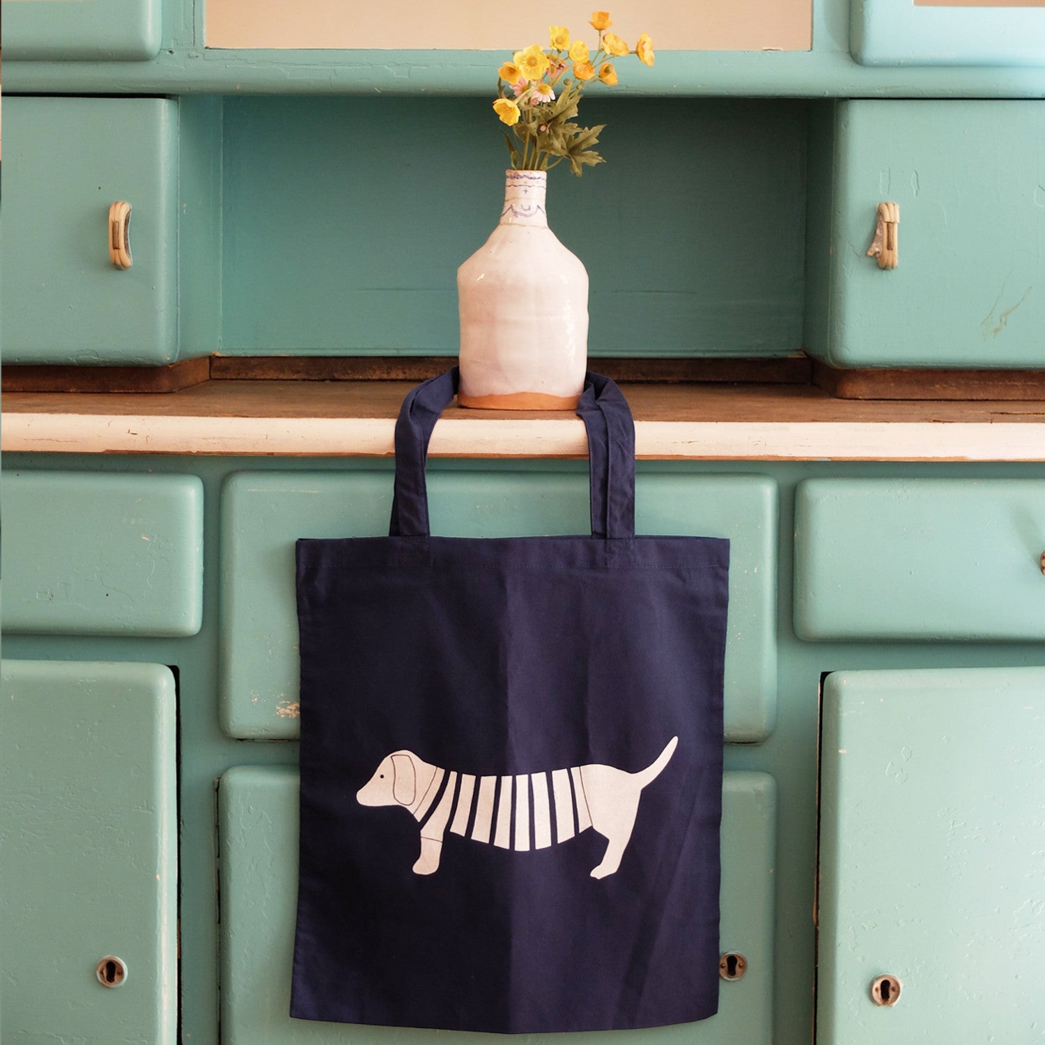 Blaue Stofftasche mit Hundemotiv hängt an einer antiken, grünen Kommode; darauf eine Vase mit gelben Blumen.