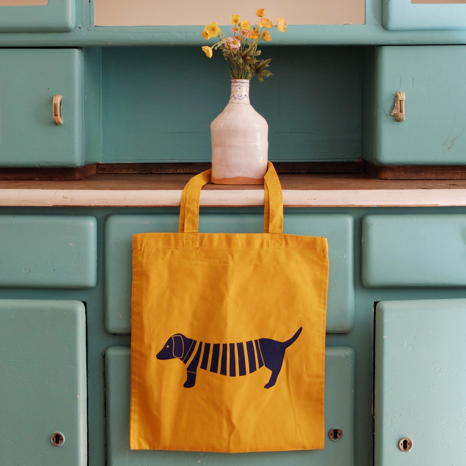Gelbe Stofftasche mit Hundemotiv hängt an einer grünen Küche; eine Vase mit gelben Blumen steht auf der Arbeitsplatte.