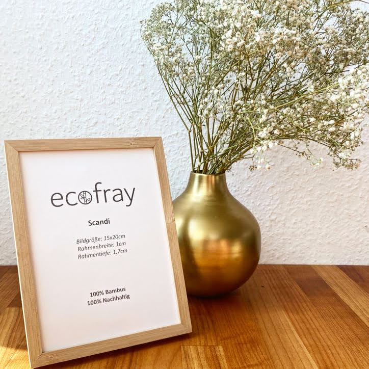 Holzrahmen mit "ecofray" Text, daneben bronzene Vase mit weißen Blüten auf Holztisch vor weißer Wand.